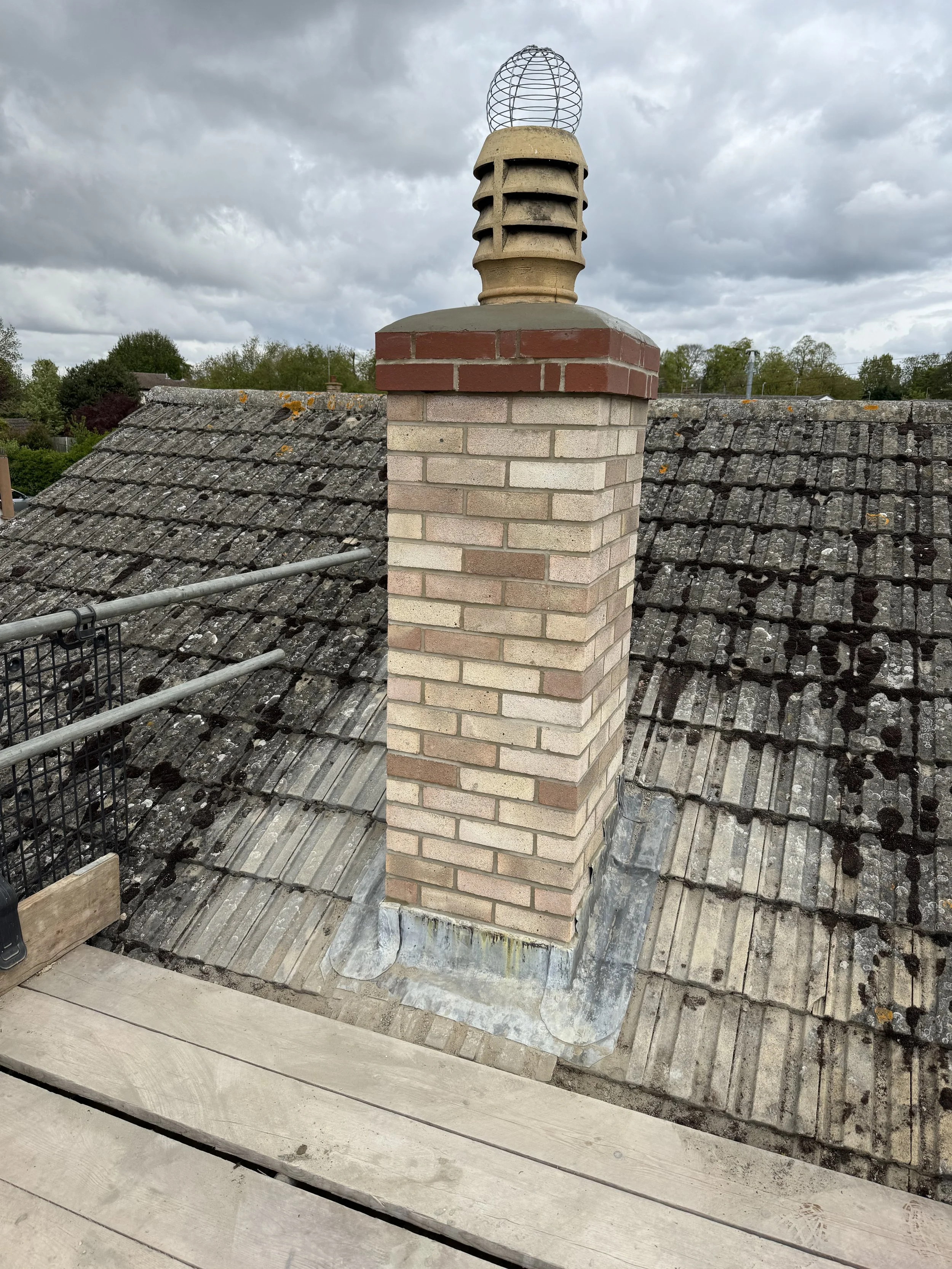 A chimney with a brick base and a metal cap is mounted on a roof covered with aged, mossy tiles. The sky is cloudy, and trees are visible in the background.