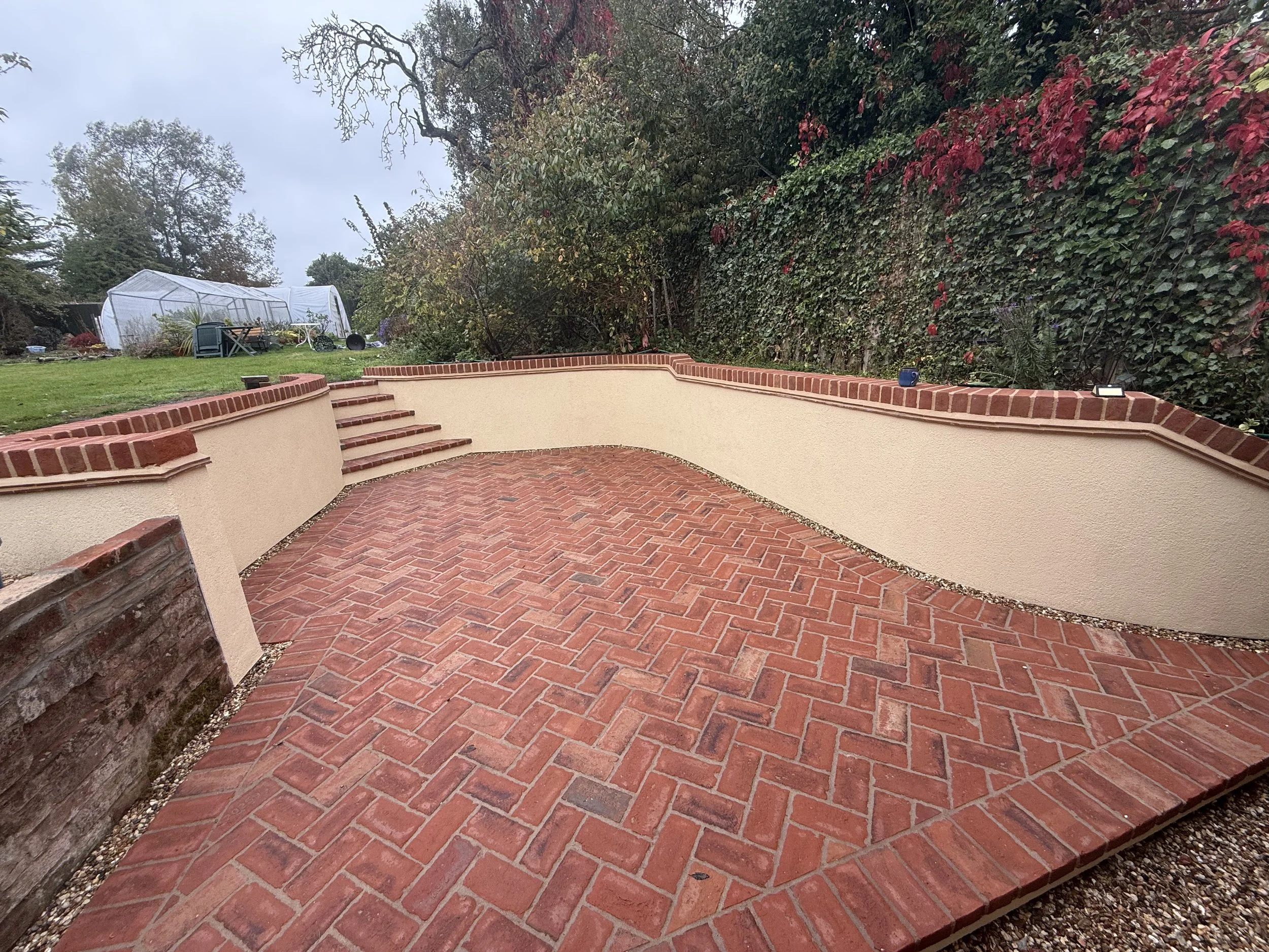 A newly constructed brick patio with curved beige stucco walls and steps, surrounded by a garden with grass, trees, and a greenhouse in the background.