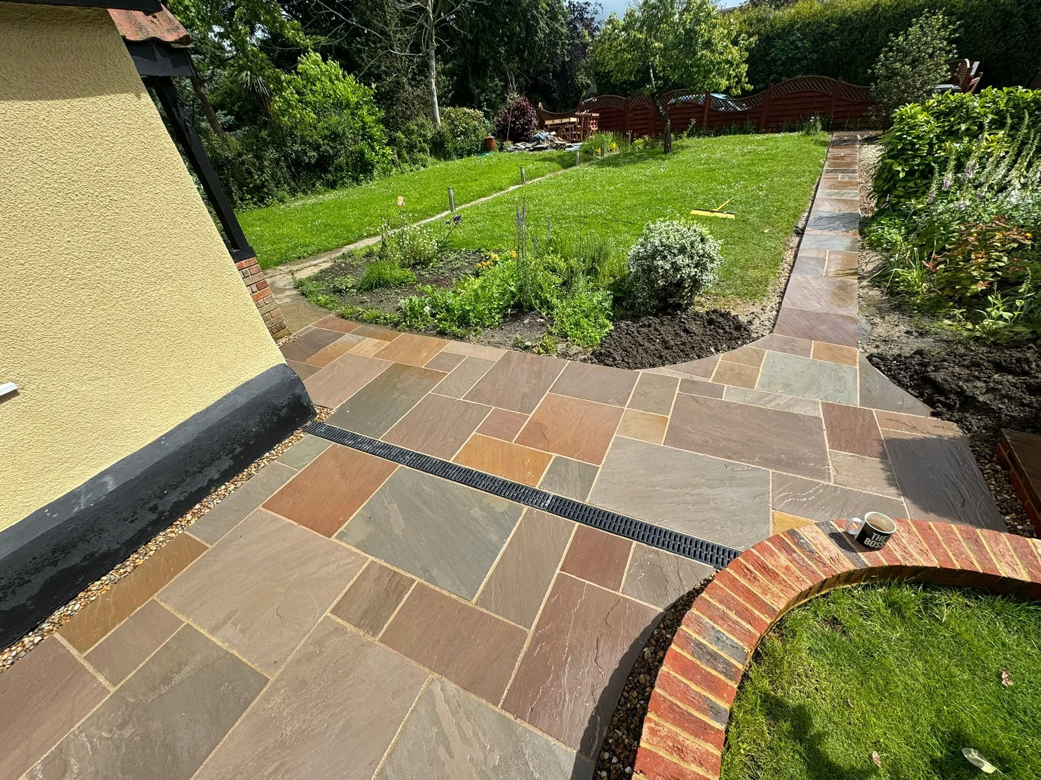 View of a well-maintained garden with a paved stone patio, green grass, shrubs, and a red wooden fence in the background.