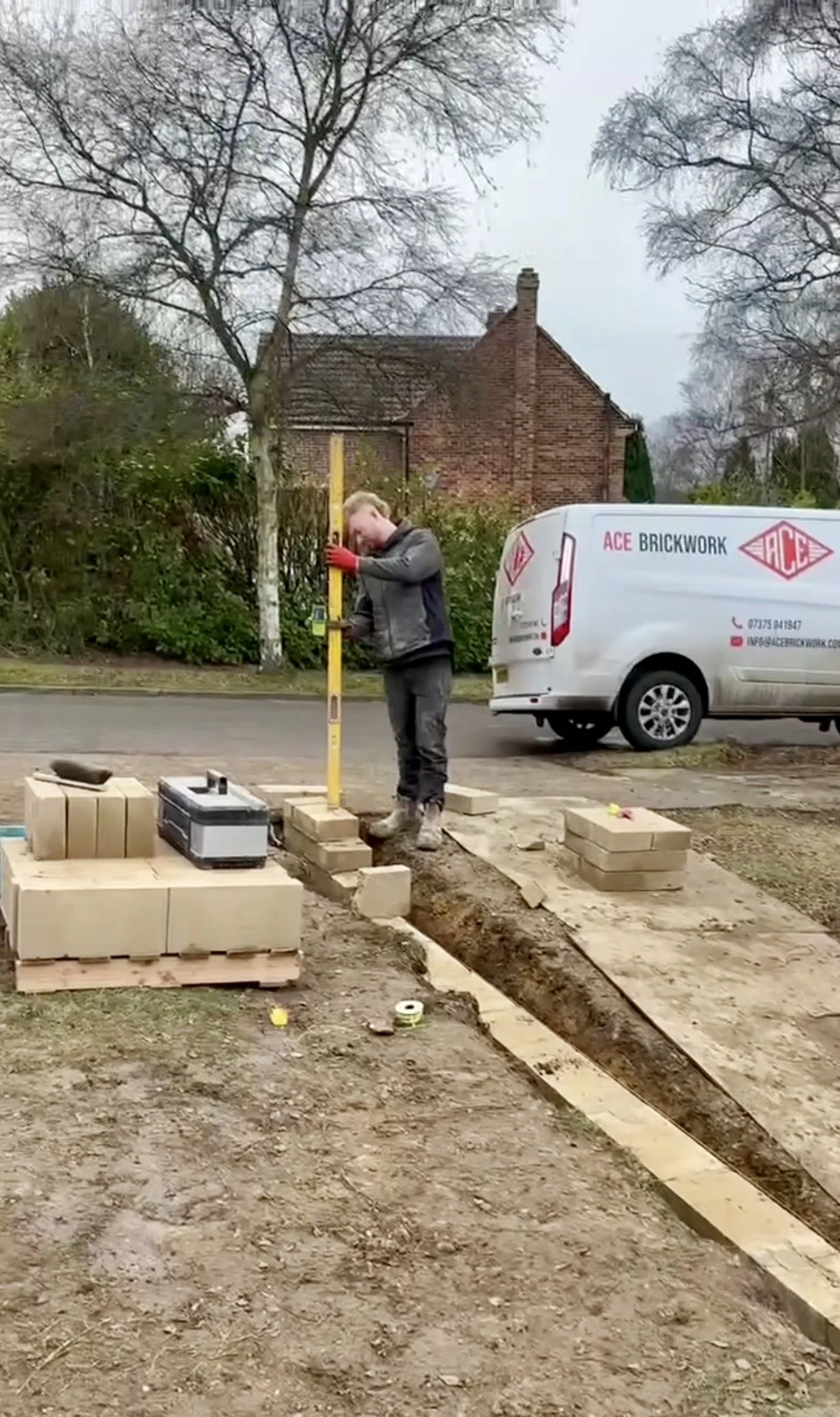 Person measuring the height of a tree with a yellow measuring stick on a construction site, with bricks, tools, and a white van labeled 'ACE BRICKWORK' in the background.
