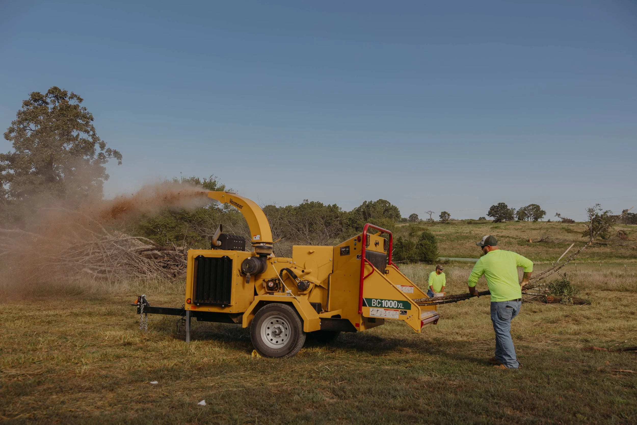 Two workers in green shirts operate a wood chipper on an open field, chipping a large tree branch into sawdust, with a background of trees and a clear blue sky.