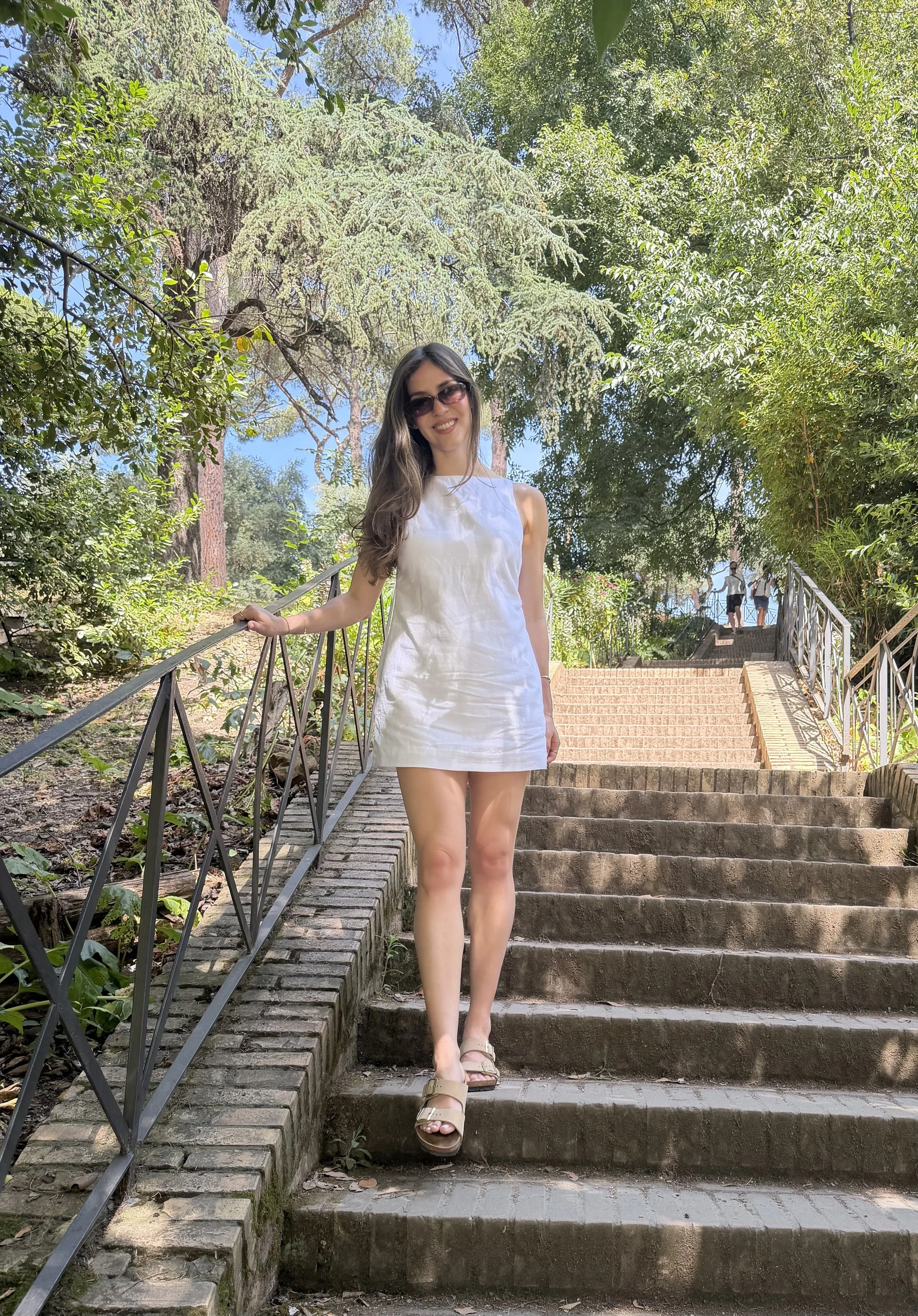 A woman with long dark hair wearing sunglasses and a white dress, standing on outdoor steps, surrounded by trees and greenery on a sunny day.