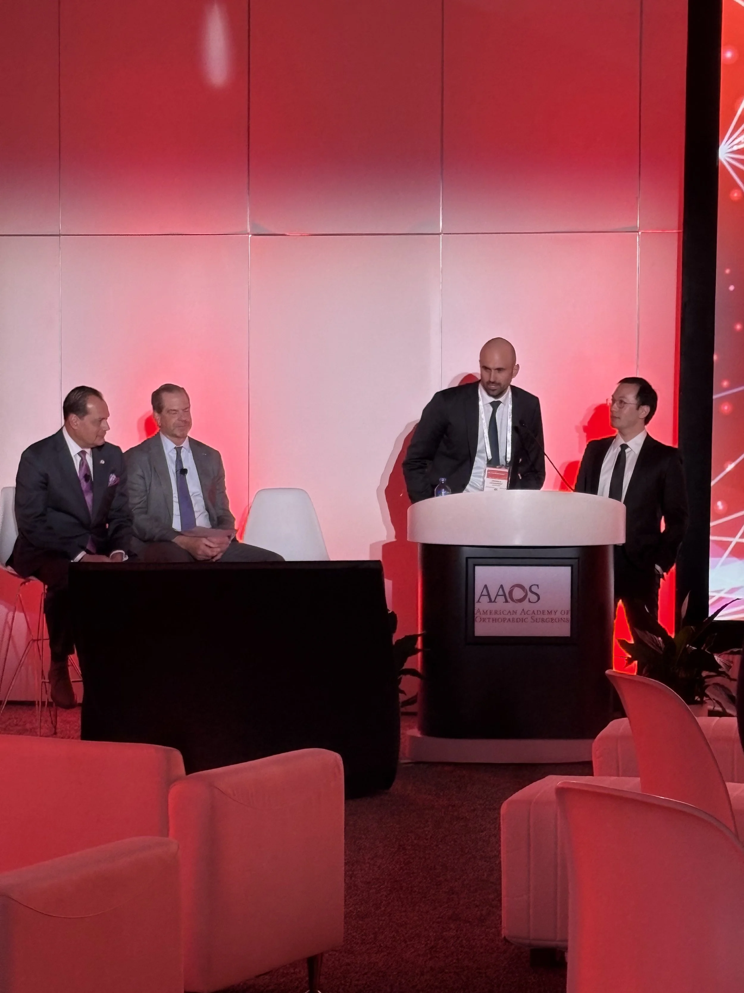 Four men in suits at a conference, two seated and two standing at a podium with a sign reading 'American Academy of Orthopaedic Surgeons.'