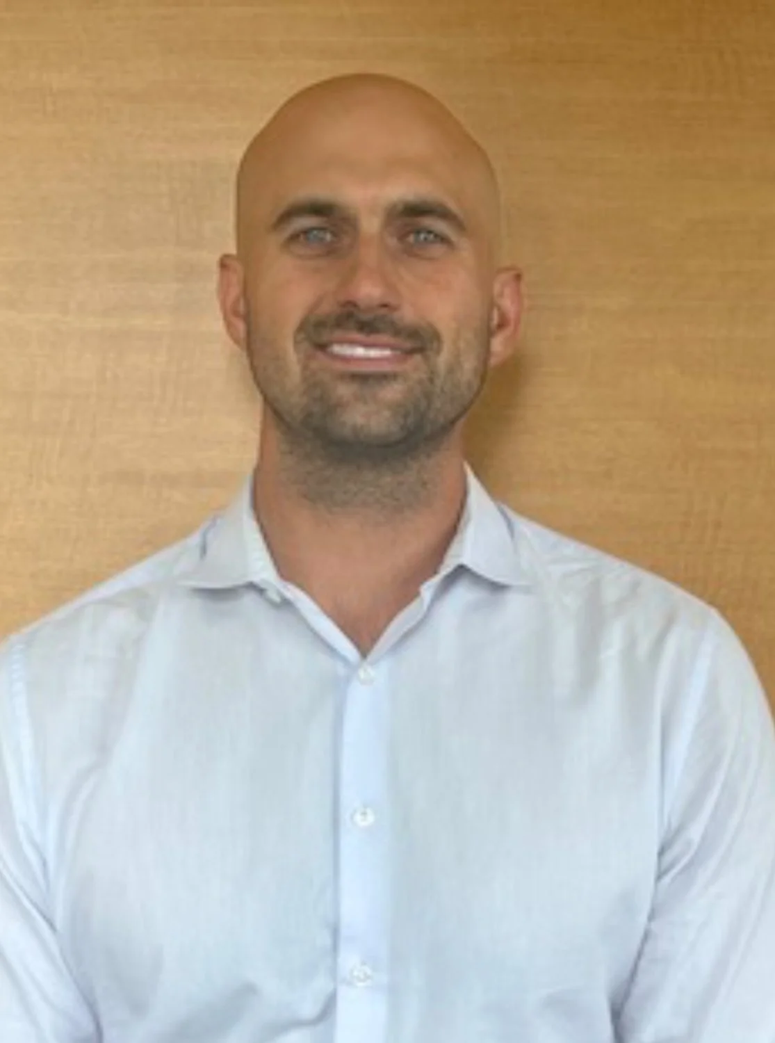 A man with a shaved head and blue eyes, wearing a white dress shirt, smiling, standing against a wood-paneled background.