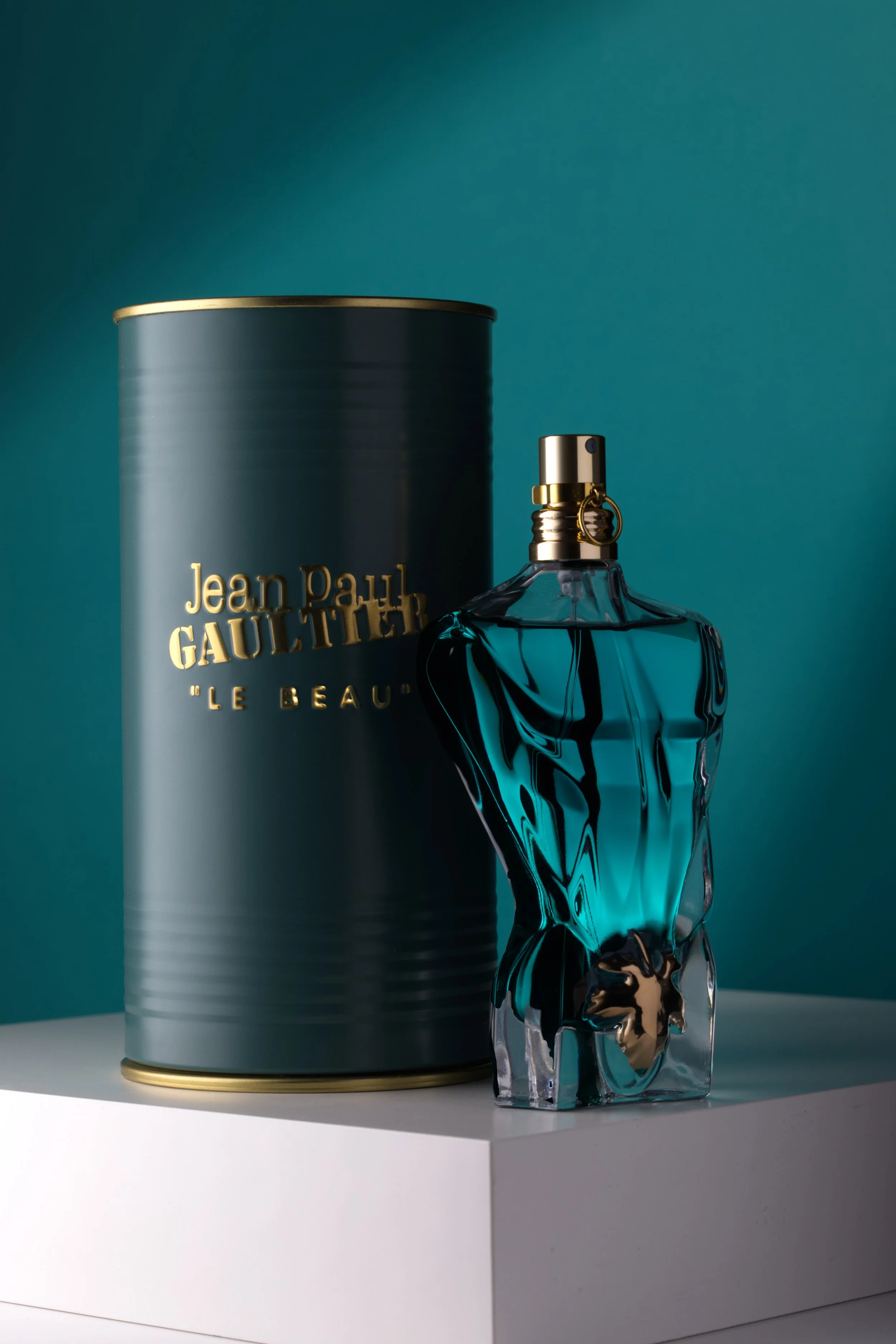 Jean Paul Gaultier "Le Beau" parfym fotograferad på vit upphöjd bas mot en grön/blå delvis belyst bakgrund.