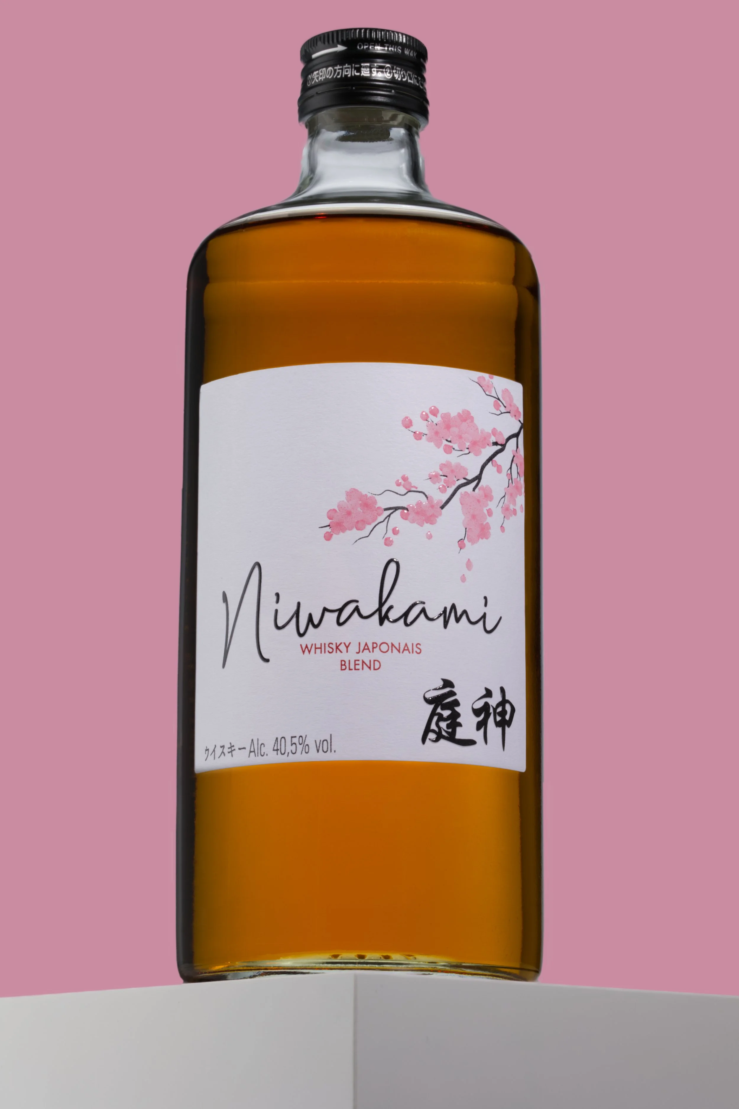 En flaska Niwakami, japansk whisky, med en vit etikett med en bild av en kvist från ett körsbärsträd/körsbärsblommor och svarta japanska tecken. Står på kanten av ett vitt bord med bakgrund i ljusrosa.