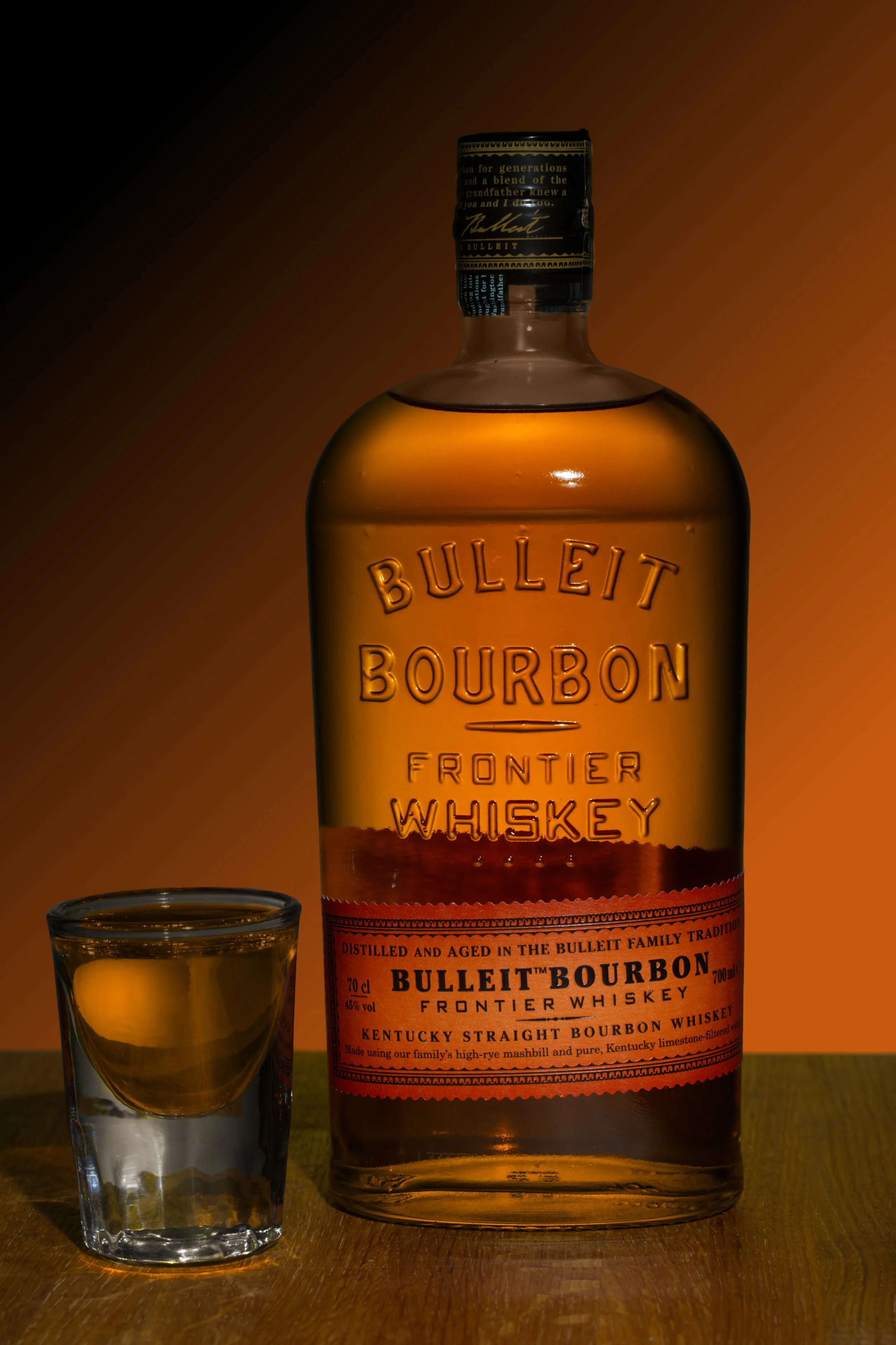 En flaska Bulleit Bourbon placerad på ett träbord med ett litet shotglas med bourbon vid sidan. Orange bakgrund som matchar den orange etiketten med svart text.
