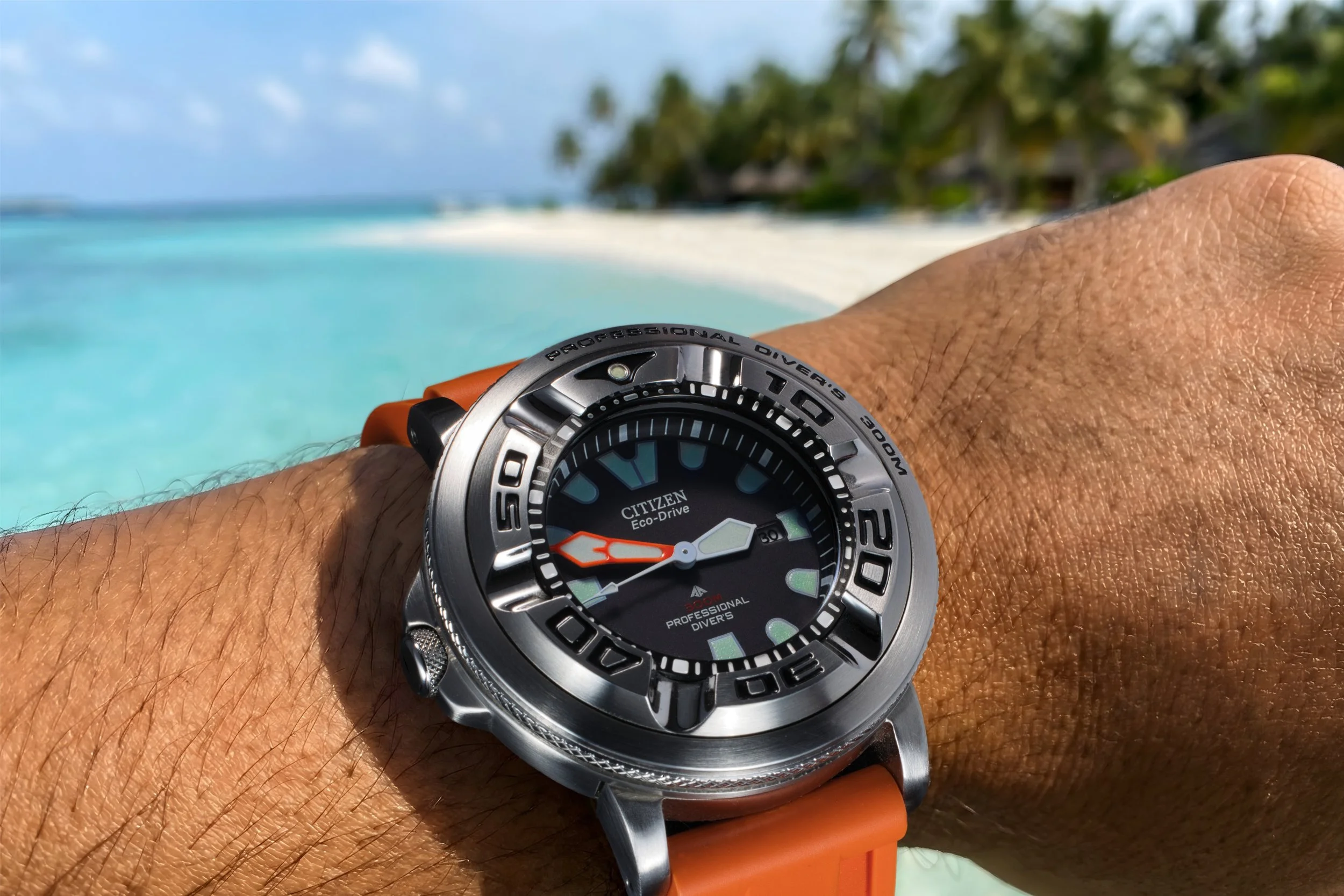 Citizen Ecozilla med orange armband på en solbränd handled i förgrunden. I bakgrunden turkost vatten och en lång sandstrand med palmer.