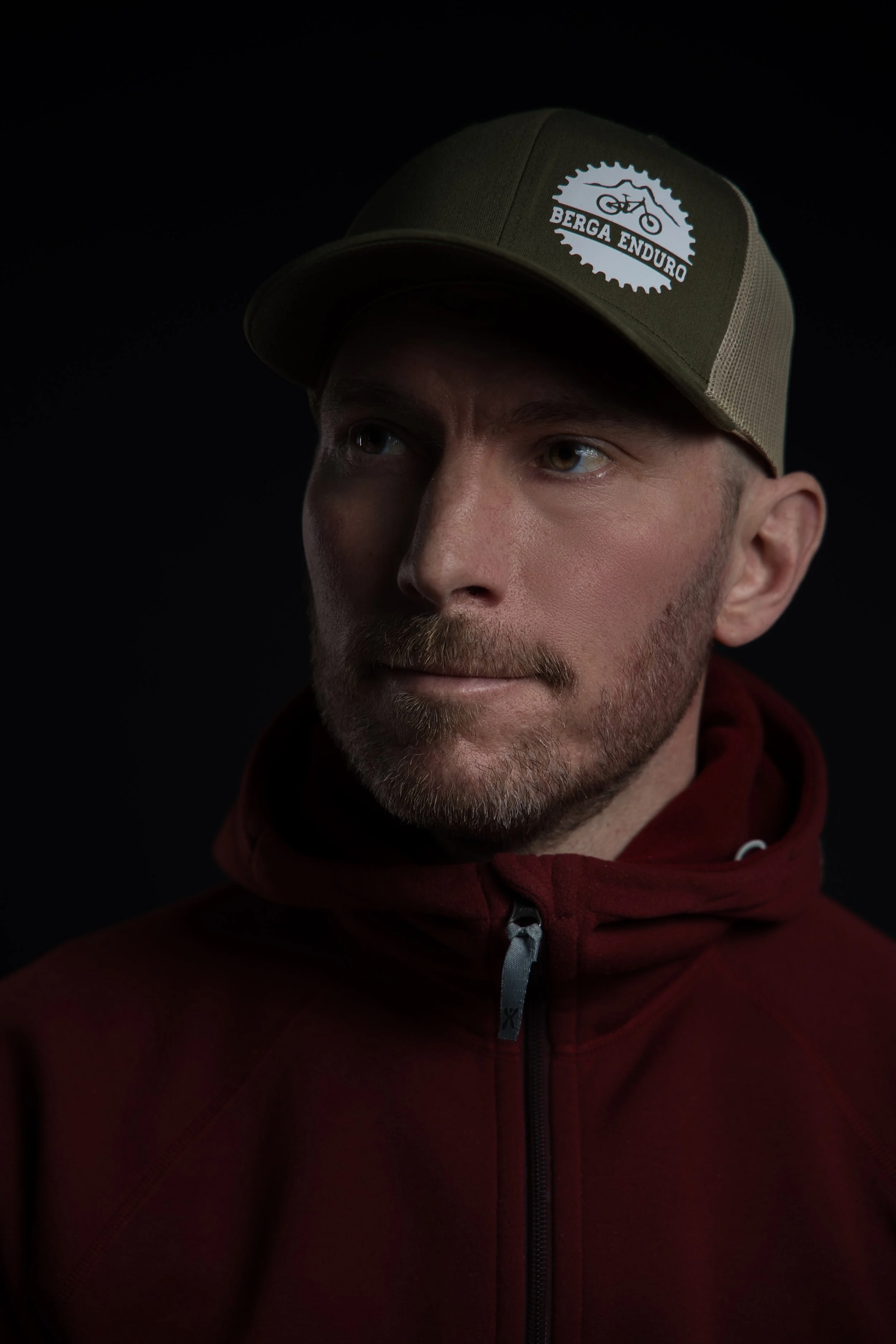 Porträttfotografi av en man med skägg, en vinröd Houdini hoodie och en grön keps som tittar åt sidan fotad mot en svart bakgrund.