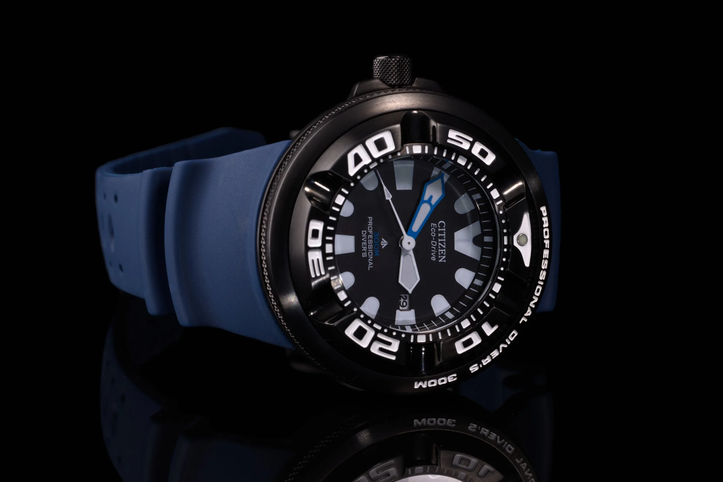 Ett Citizen Eco-Drive ("Ecozilla") armbandsur i svart stål och med blått silikonarmband liggandes på sida med reflektion mot svart glansigt underlag och svart bakgrund.