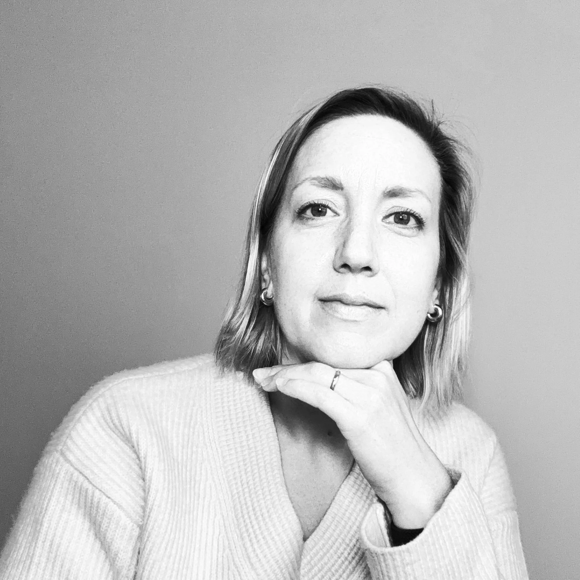 A woman with shoulder-length hair, wearing earrings and a light-colored sweater, resting her chin on her hand in a black-and-white portrait.