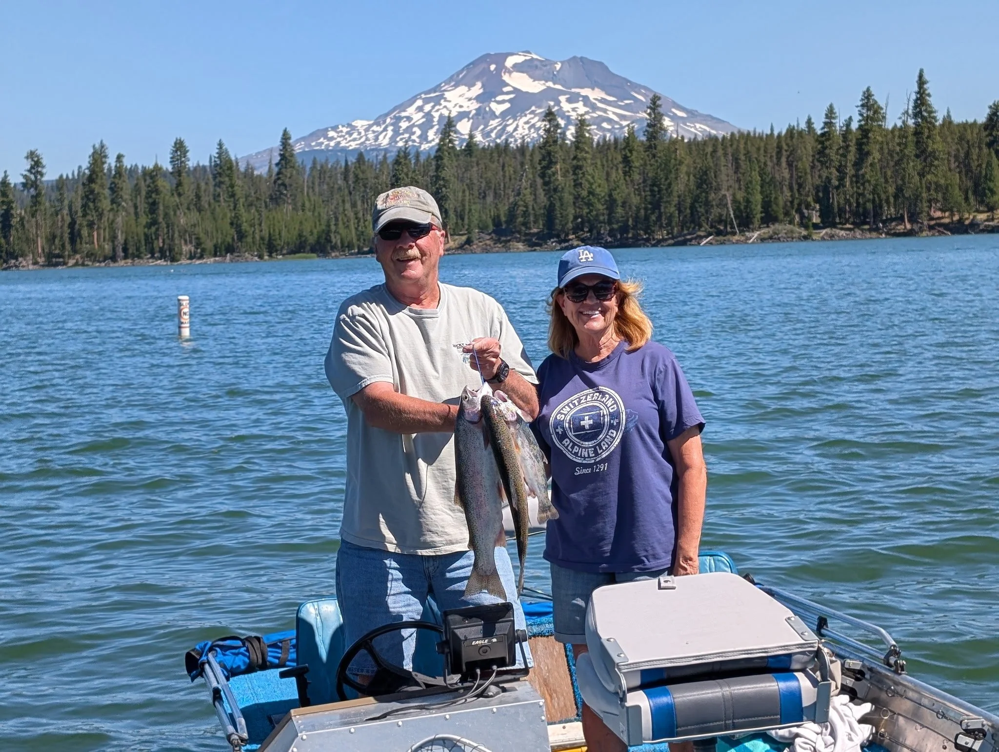 A man and a woman on a boat holding a large fish, with a mountain and lake in the background.