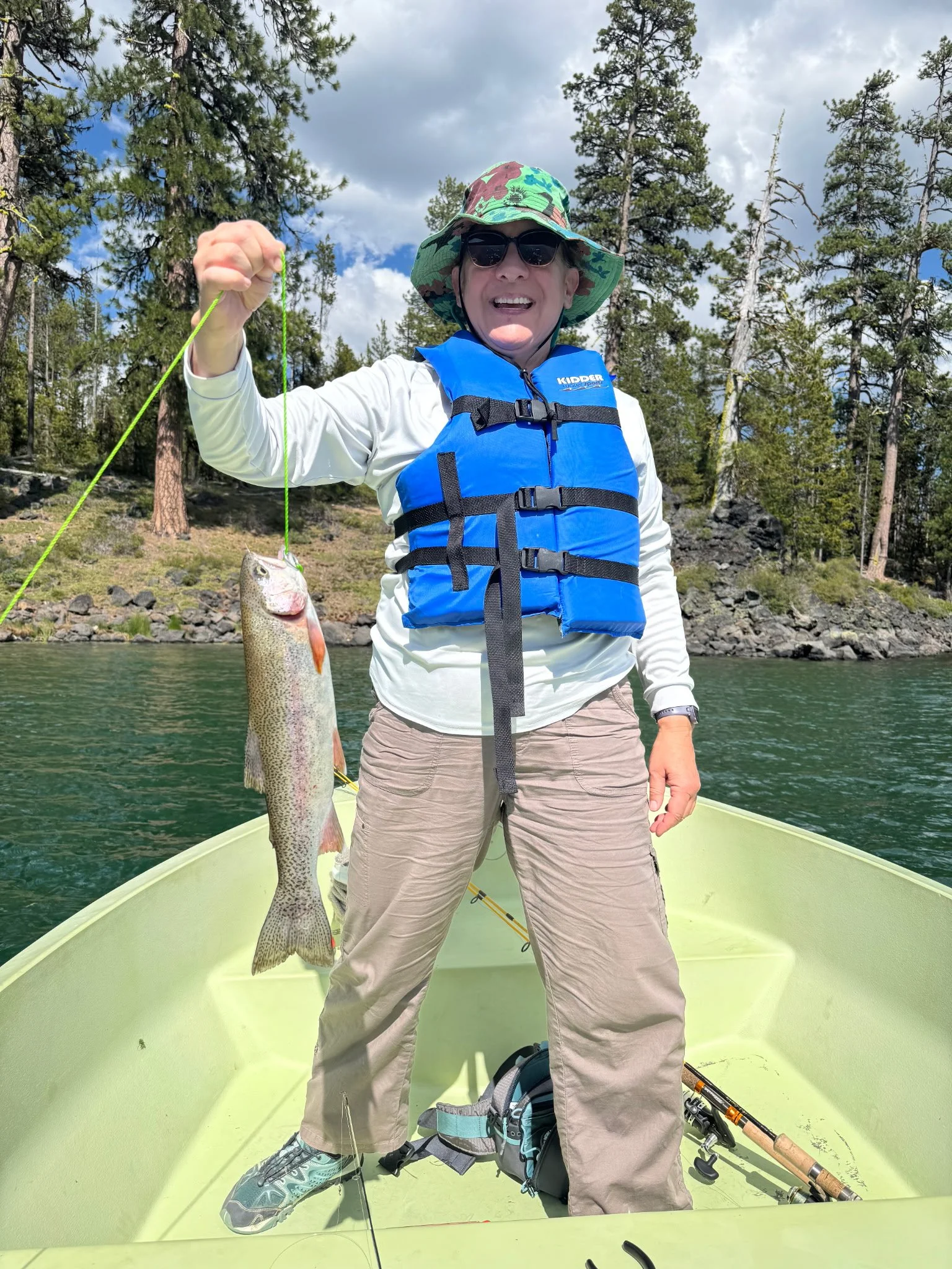 A person standing in a boat holding a fish they caught, wearing sunglasses, a wide-brimmed hat, a blue life vest, and casual outdoor clothing, with a forest and cloudy sky in the background.