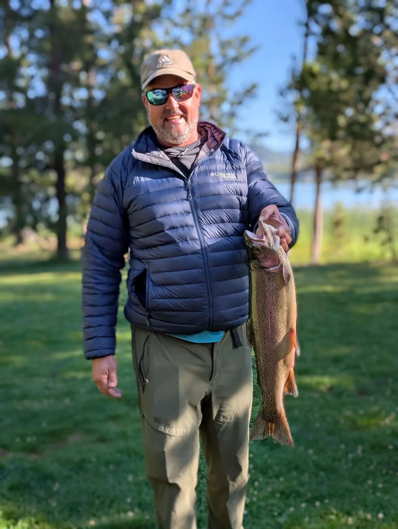 A man outdoors holding a large fish, standing on grass near trees and a body of water.