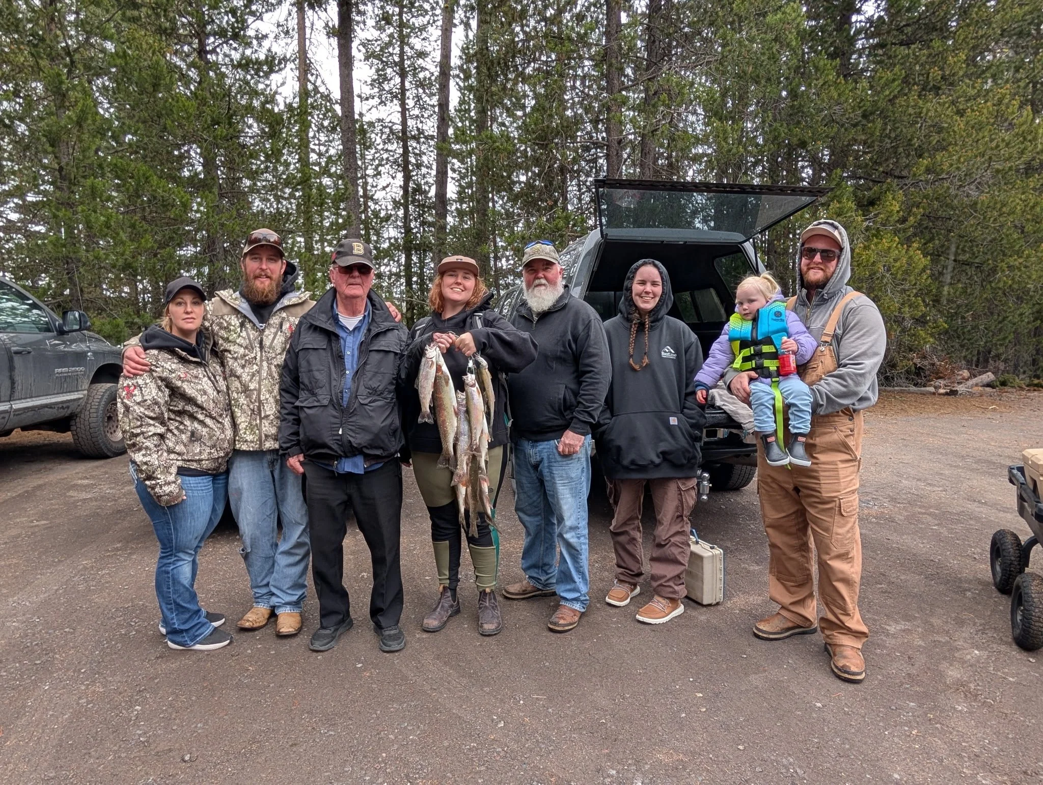 Group of nine people standing outdoors in a forested area, holding fishing gear and fish, with a black SUV and a pickup truck nearby, celebrating a fishing trip.