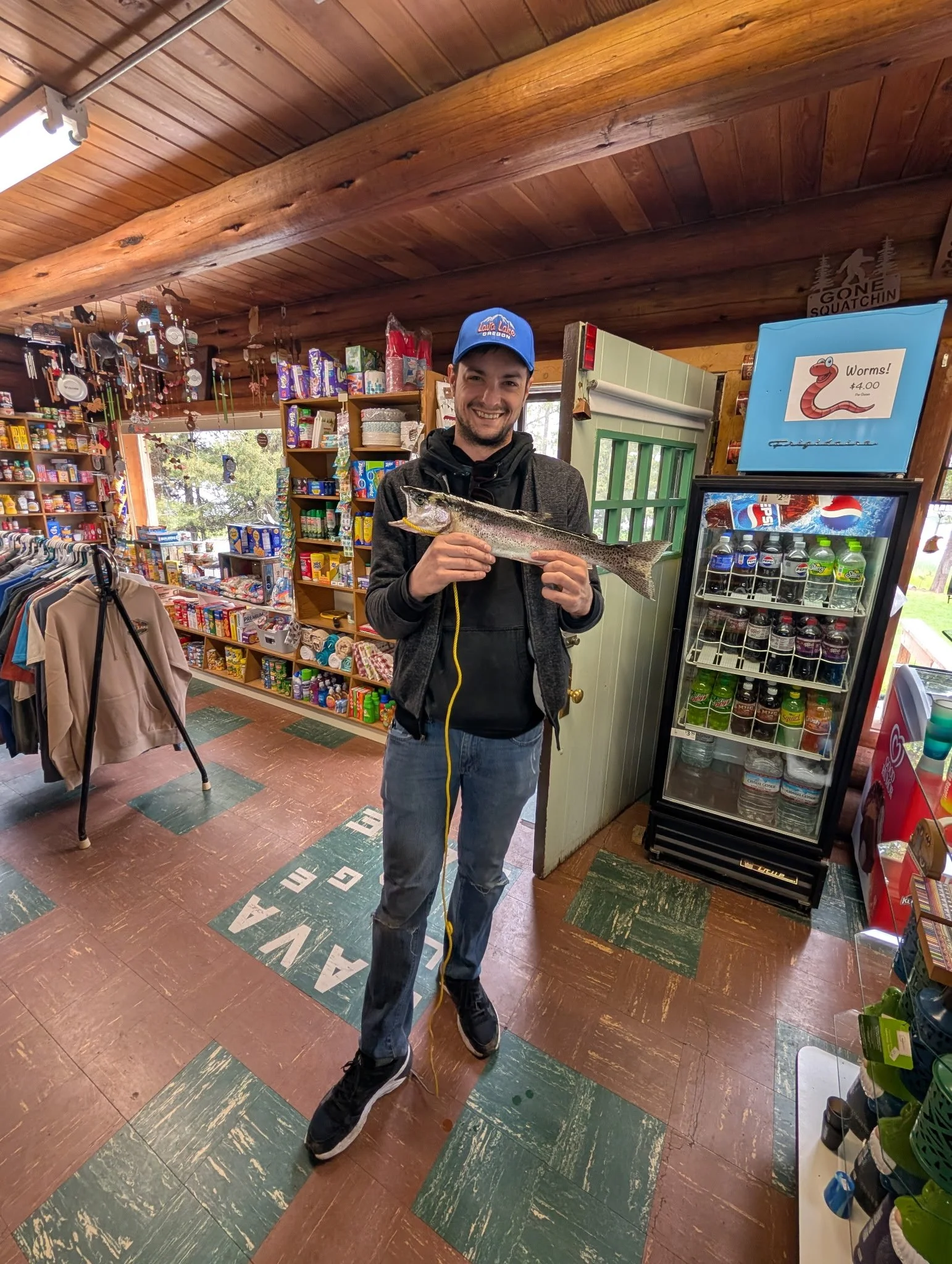 A smiling man holding a large fish indoors in a store, surrounded by snacks and drinks.