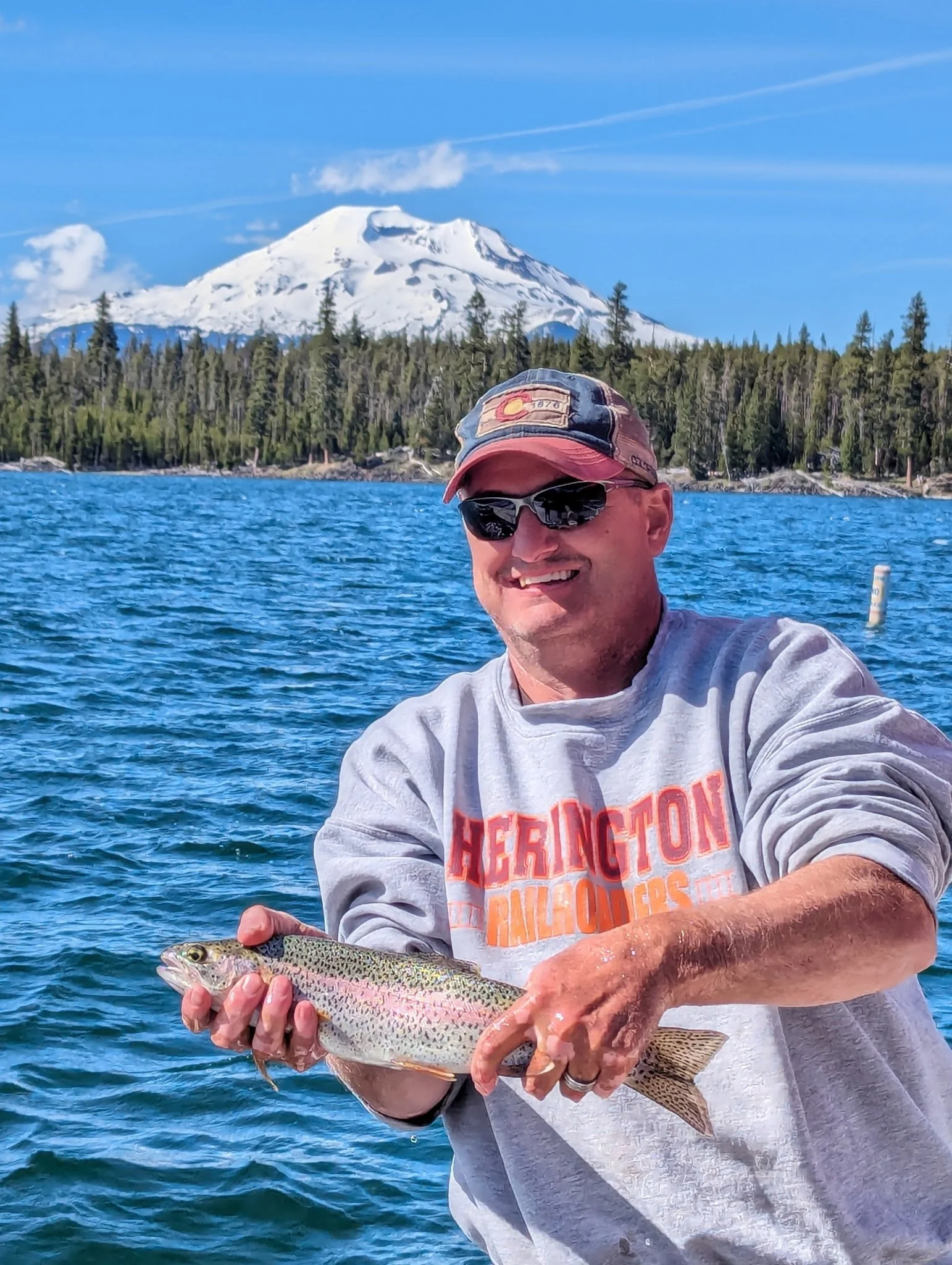 Man smiling while holding a rainbow trout in a lake, with a snow-capped mountain and forested shoreline in the background on a clear day.