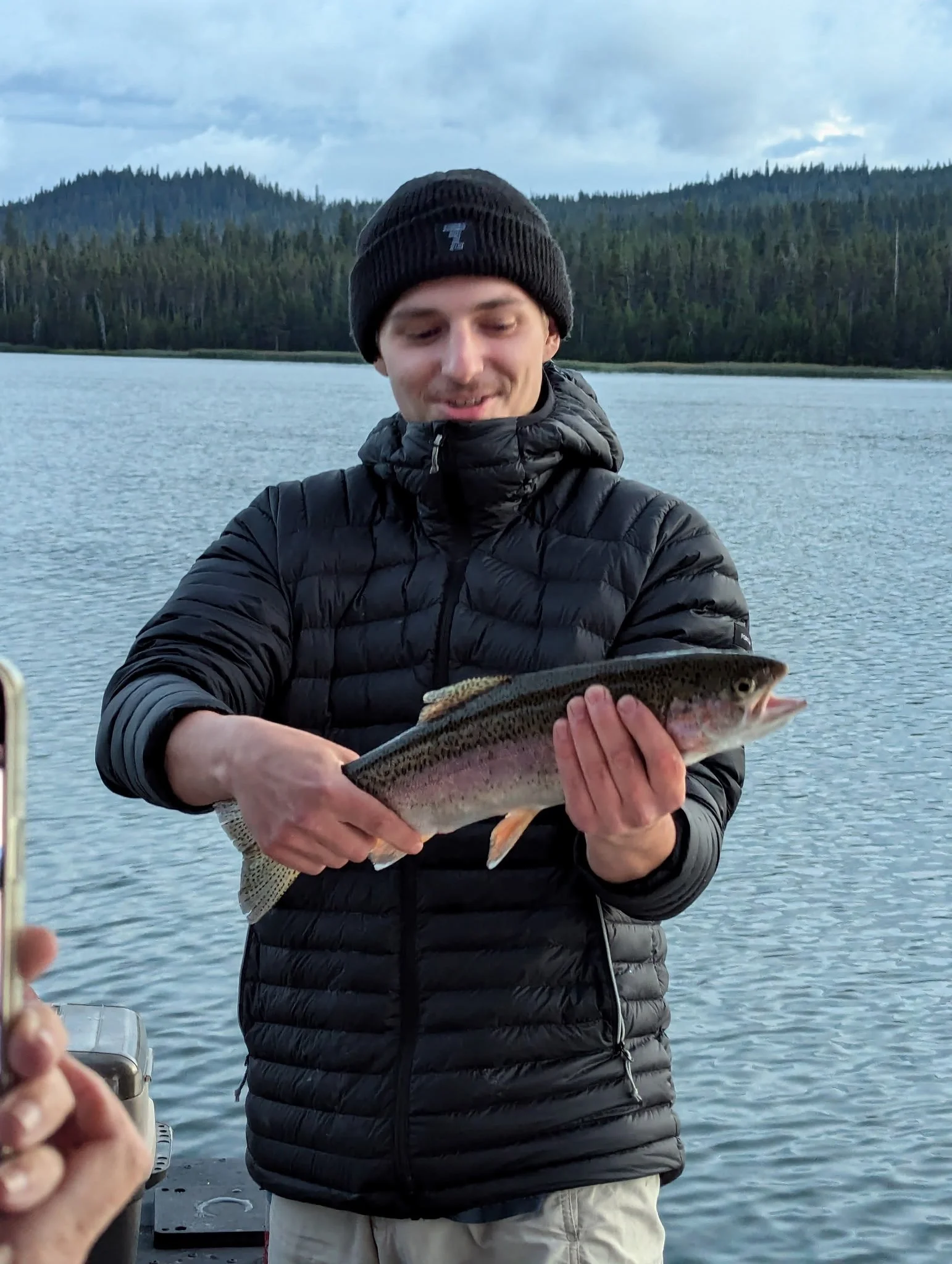 A young man in a black beanie and puffy jacket holding a rainbow trout near a lake, with forested hills in the background.