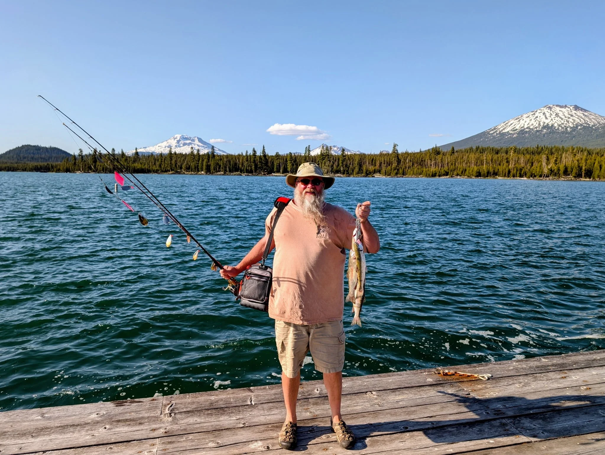 Man standing on wooden dock holding fishing rod and a fish, with lake, forest, and snow-capped mountains in the background.