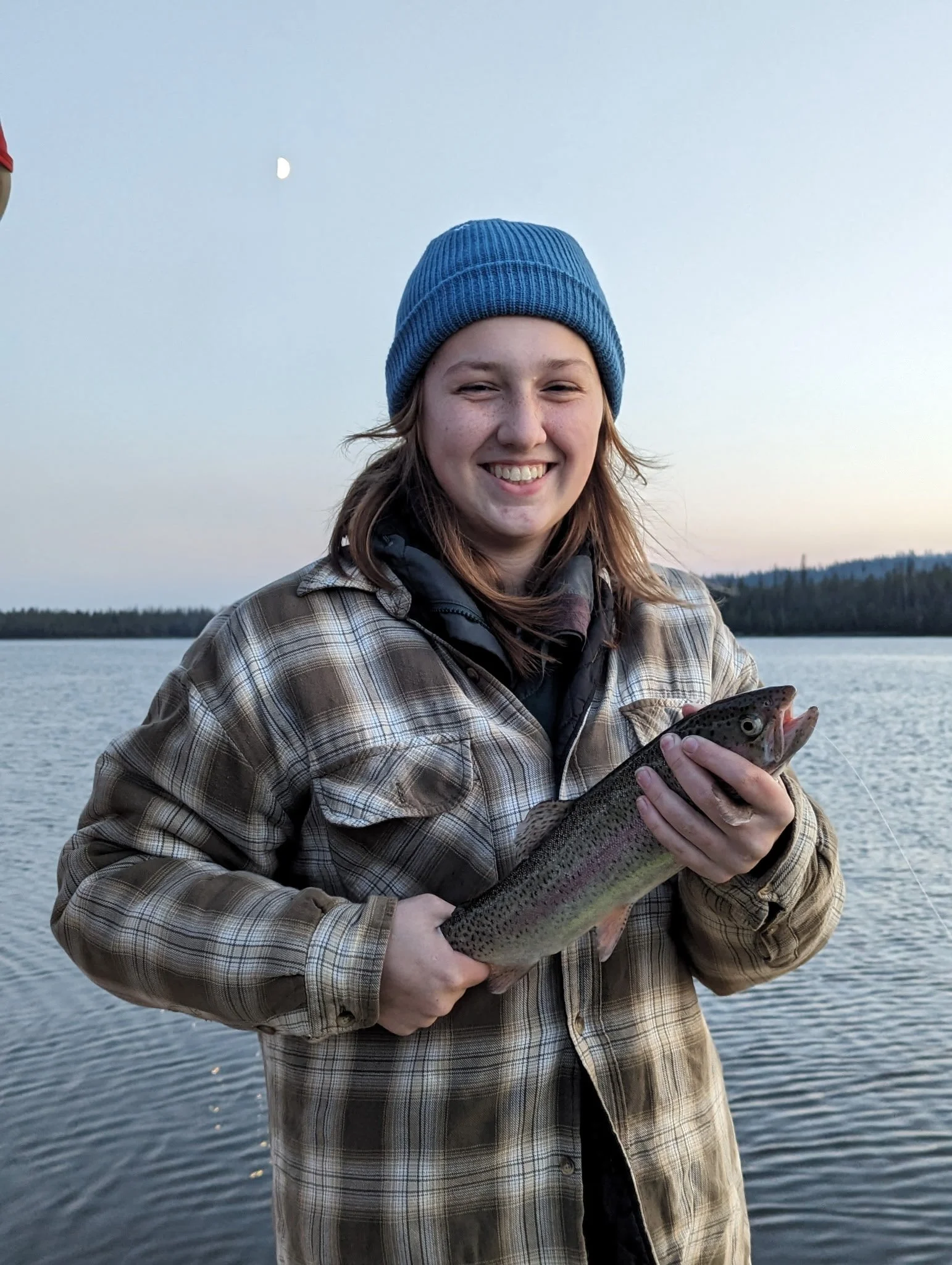 Girl in a blue beanie and plaid jacket smiling while holding a rainbow trout by a lake during dusk.
