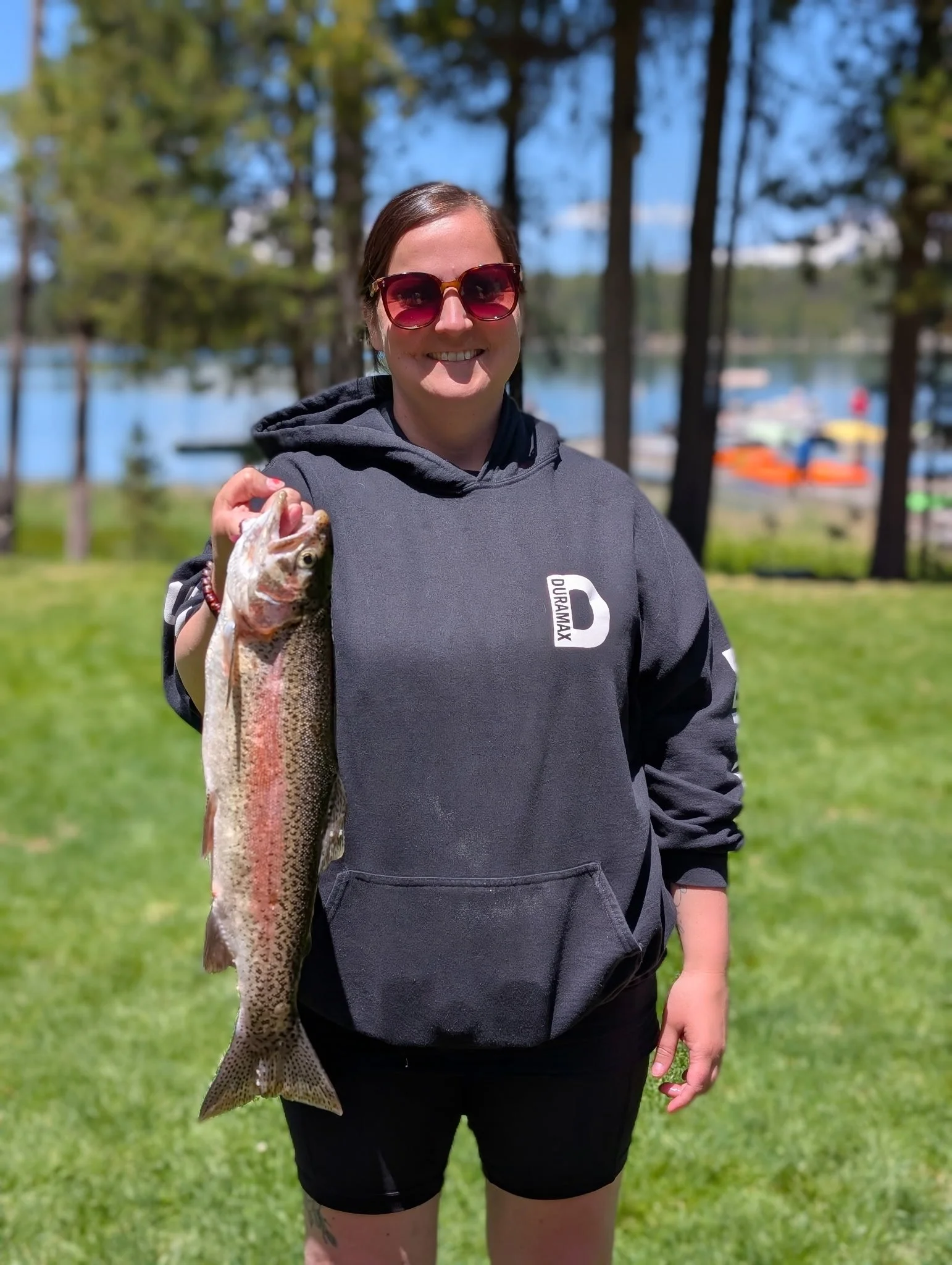 A woman wearing sunglasses and a black hoodie is standing on grass, holding a large fish she caught, with a lake and trees in the background.