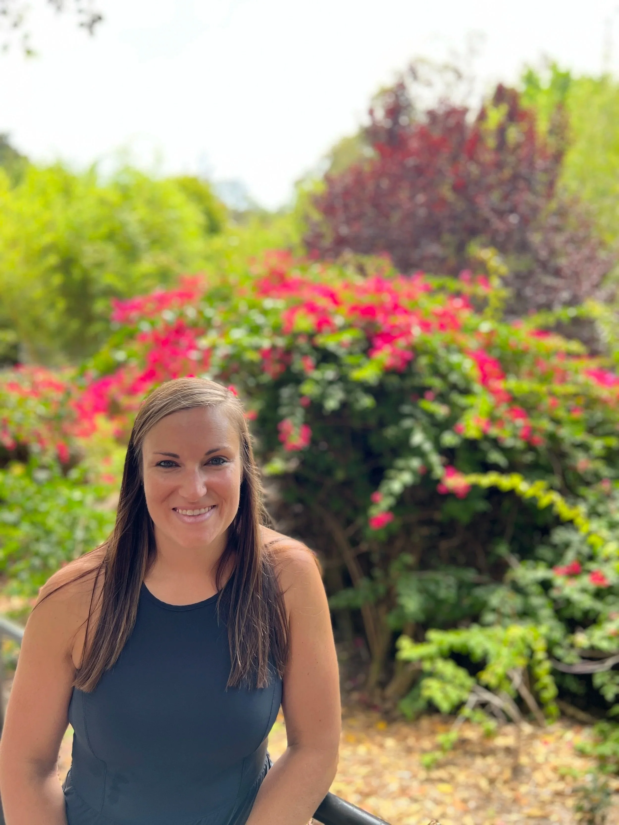A smiling woman with brown hair in a blue sleeveless top posing outdoors in front of blossoming pink flowers and green foliage.