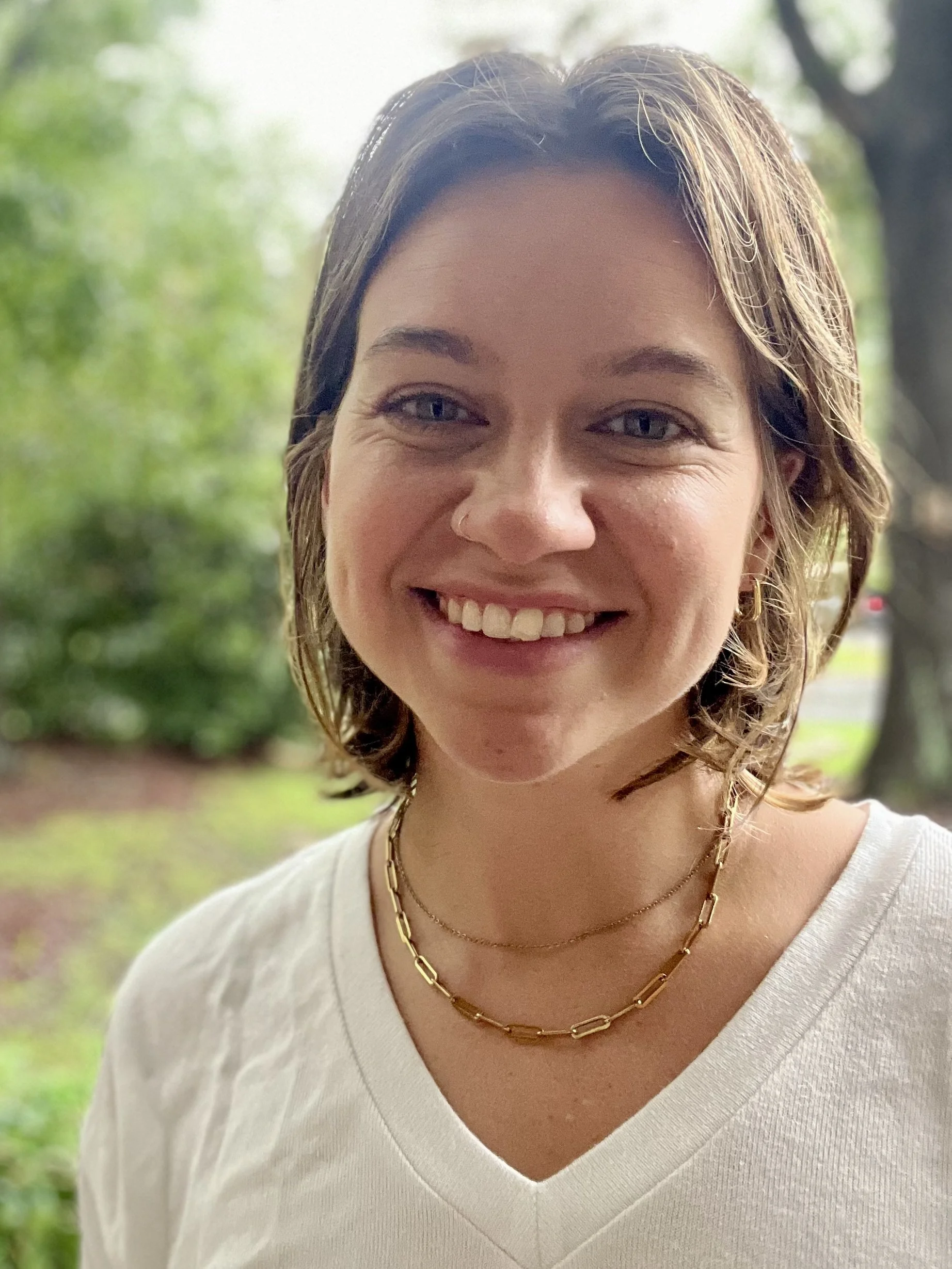 A young woman with short brown hair and blue eyes smiling outdoors with a blurred green background.