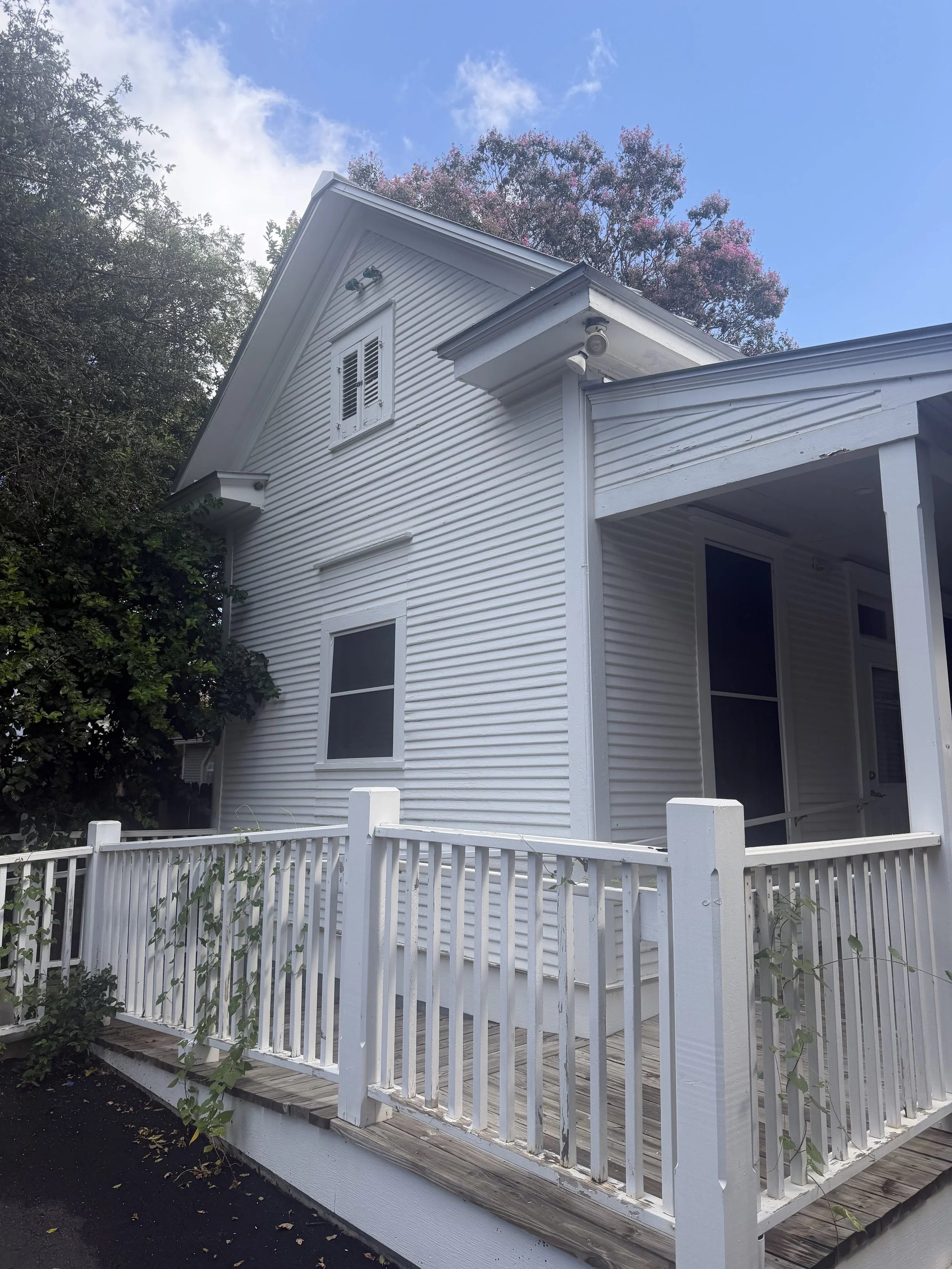 White house with a front porch, fenced yard, and a trees with pink flowers in the background sky is partly cloudy.
