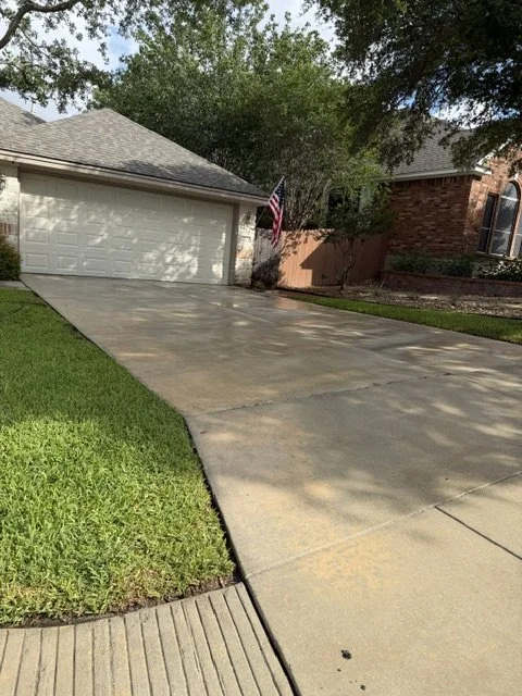 Concrete driveway leading to a garage, bordered by green grass on one side and a brick house with an American flag in the background.