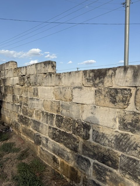 A weathered beige brick wall with blackened patches, beneath a blue sky with white clouds and power lines.