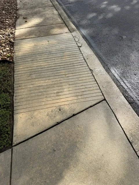 A sidewalk with textured and smooth concrete sections next to a curb and street, with a small patch of grass and gravel on the side.