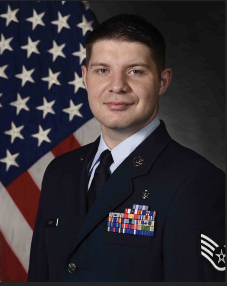 A young man in a U.S. military uniform standing in front of an American flag.