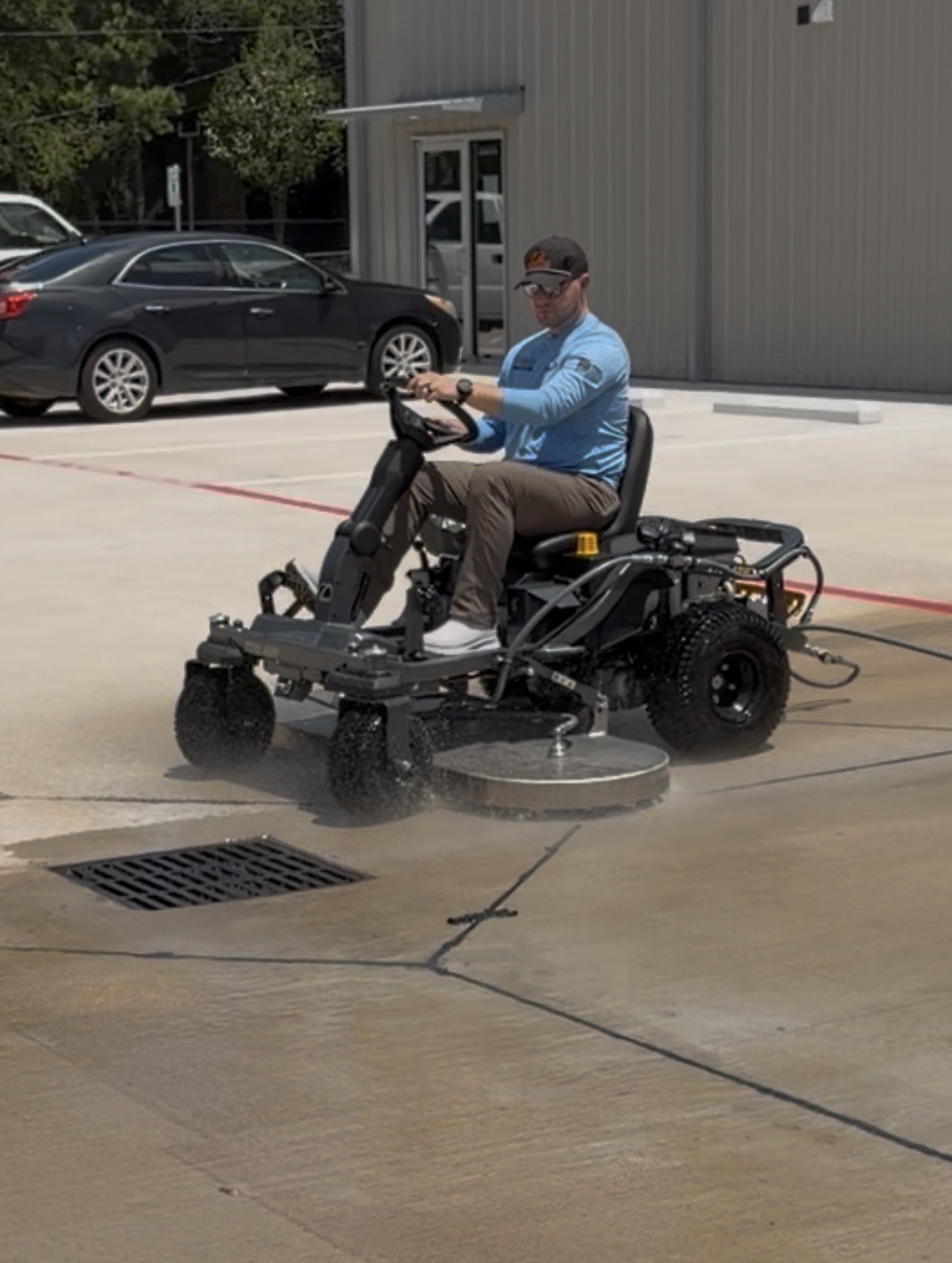 A man in a blue shirt and brown pants is operating a street cleaning machine outdoors in a parking lot. The machine has large black wheels and brushes underneath, cleaning the pavement.