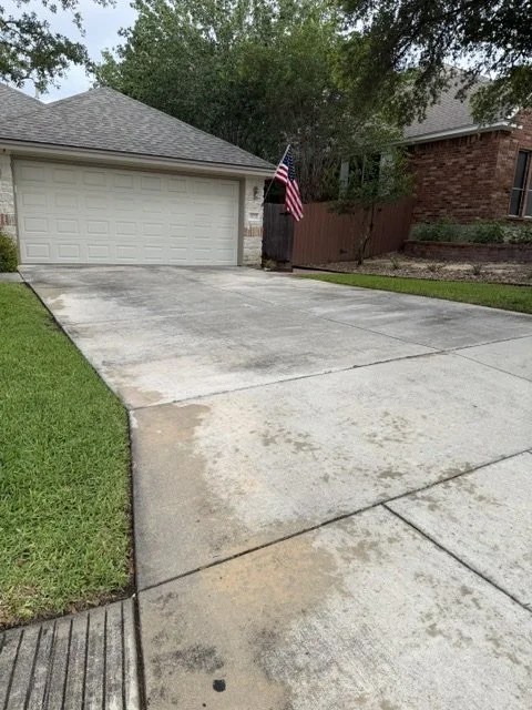 Empty driveway in front of a garage, with an American flag on a pole near a wooden fence, and a brick house partially visible to the right.