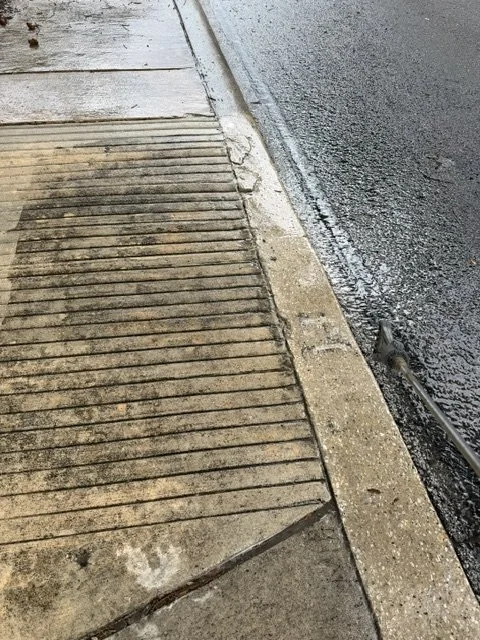 Close-up of a sidewalk and a street curb with a small drainage grate along the curb on a rainy day.