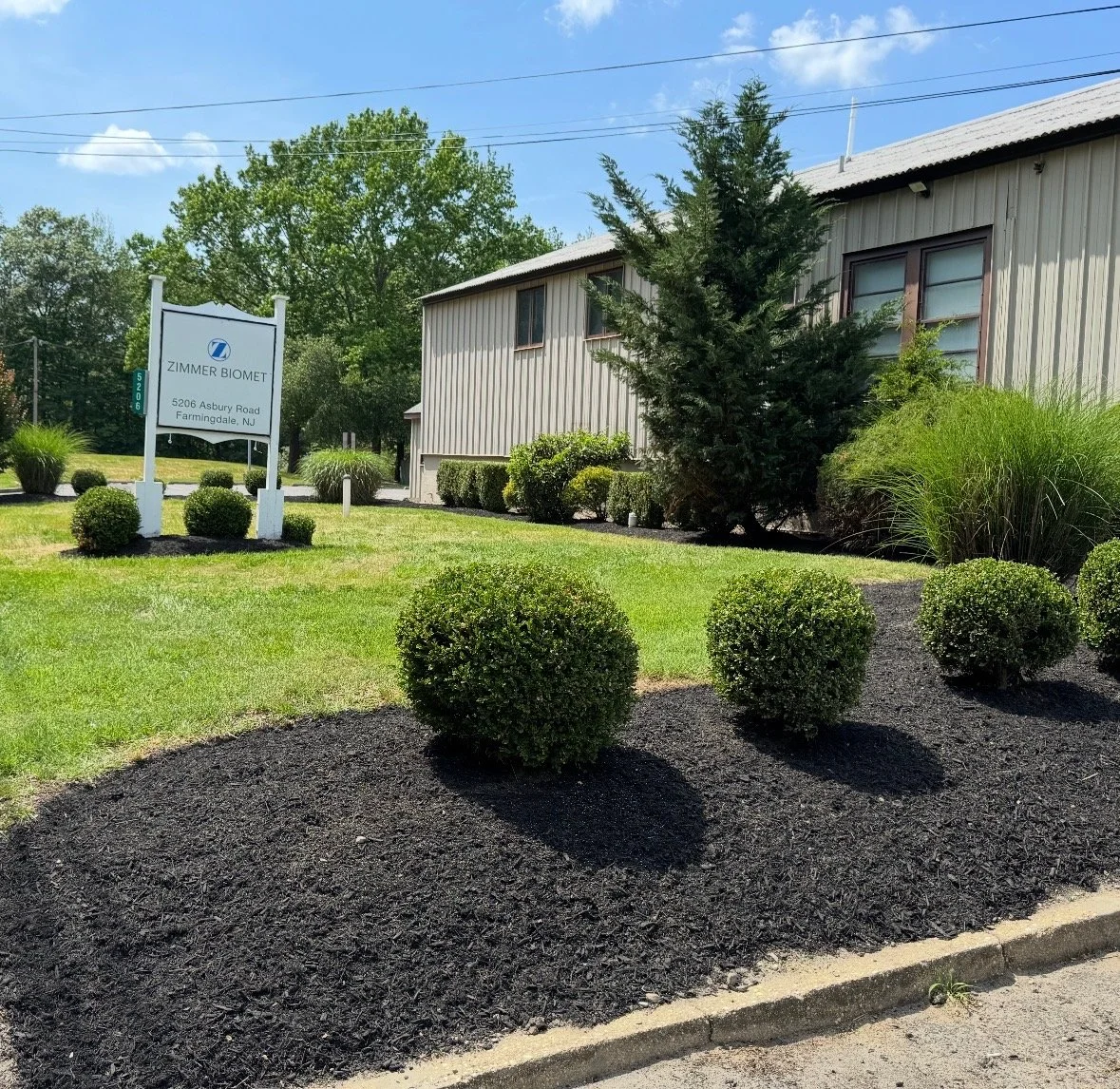The image shows a landscaped area with trimmed bushes and a small tree in front of a beige metal building. There is a white sign that reads 'Zimmer Biomet, 5206 Asbury Road, Farmingdale, NJ'. The sky is blue with a few clouds and there are power lines above.