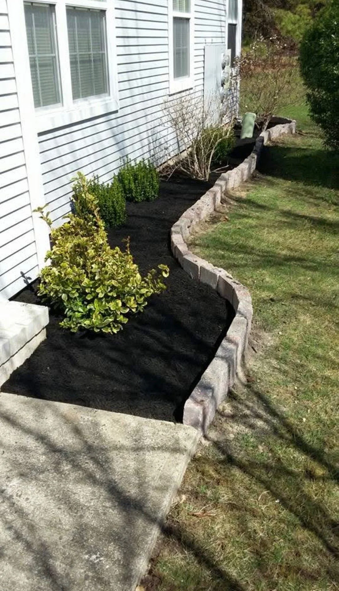 A landscaped garden bed along the side of a house with white siding, featuring plants and bushes, bordered by a stone wall and a concrete sidewalk.
