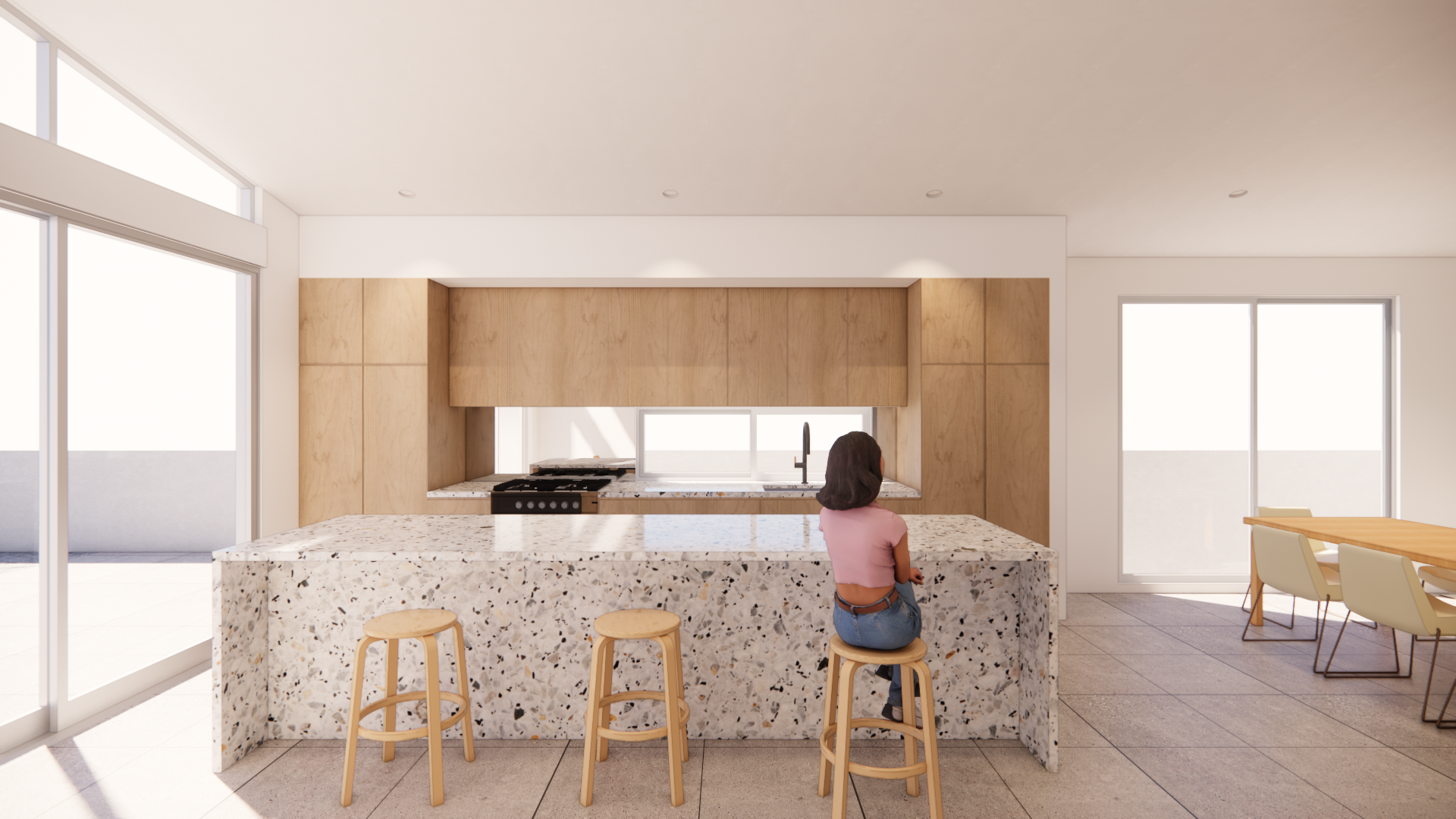 Modern kitchen with a large granite island, light wood cabinets, and a woman sitting on a stool at the island. Bright natural light from large windows and sliding doors.