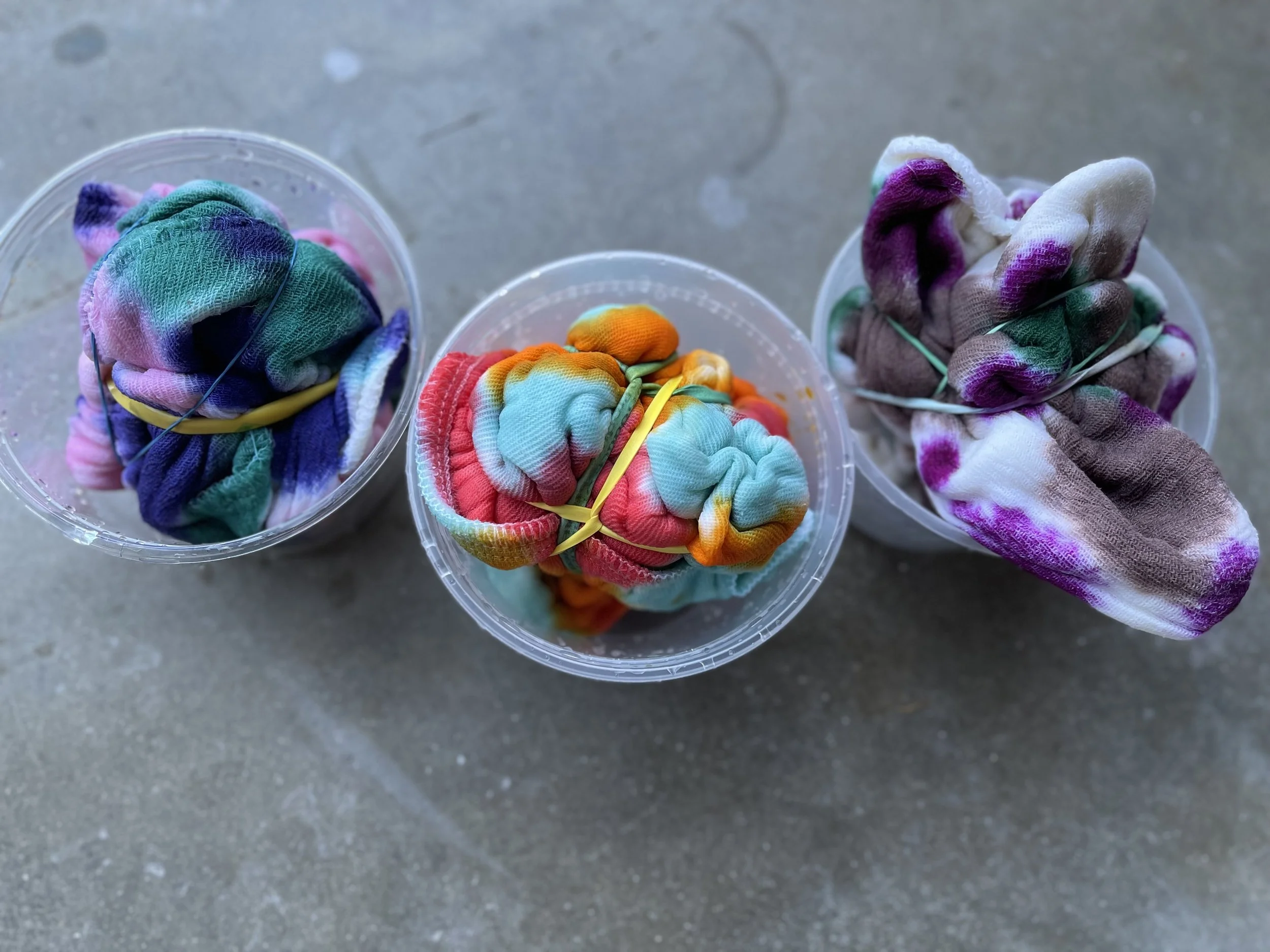 Three clear plastic cups filled with colorful tied-up cloth scraps resting on a gray surface.