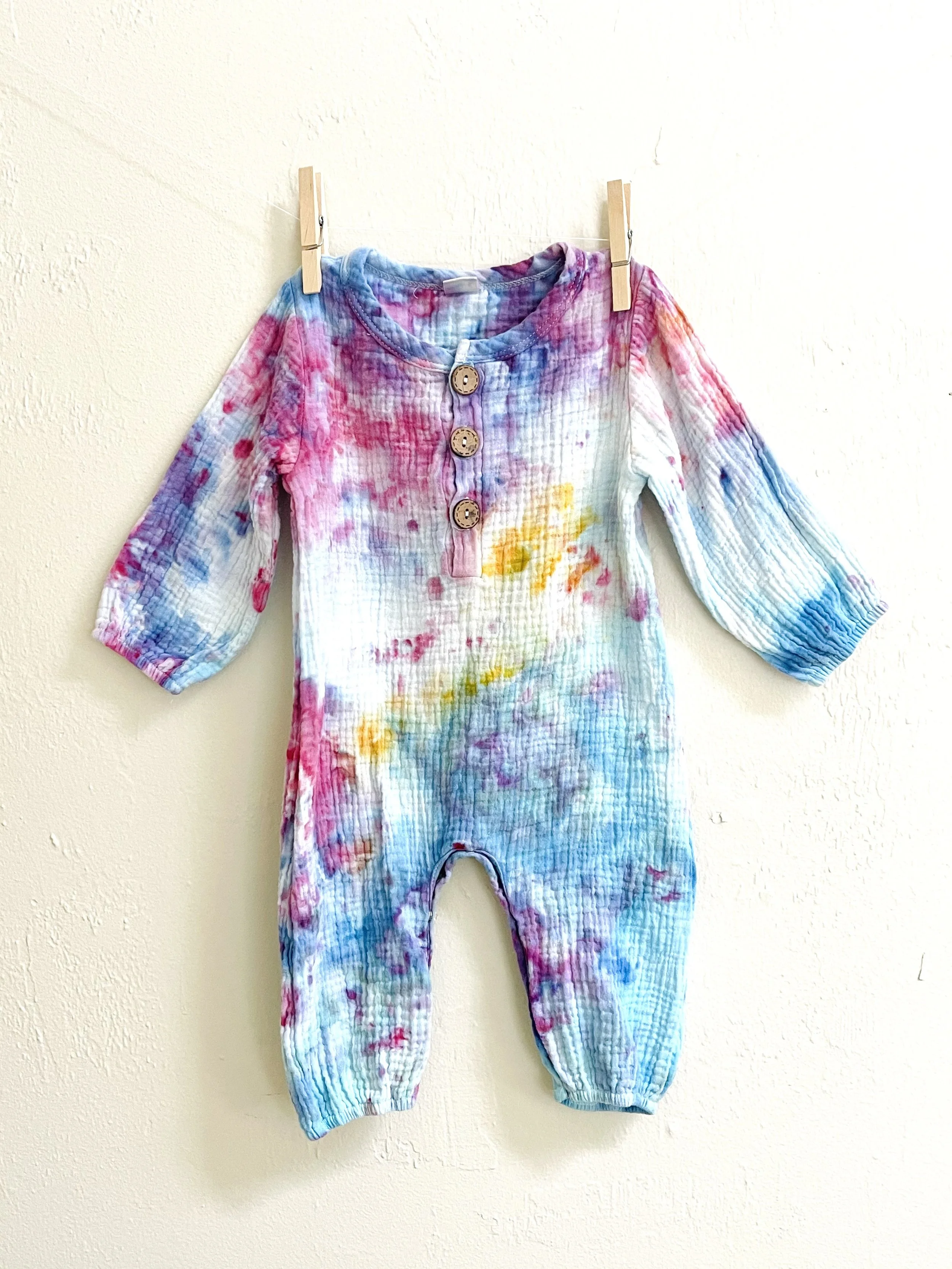 Colorful, tie-dye baby romper with three buttons on the front, hanging on a wall.
