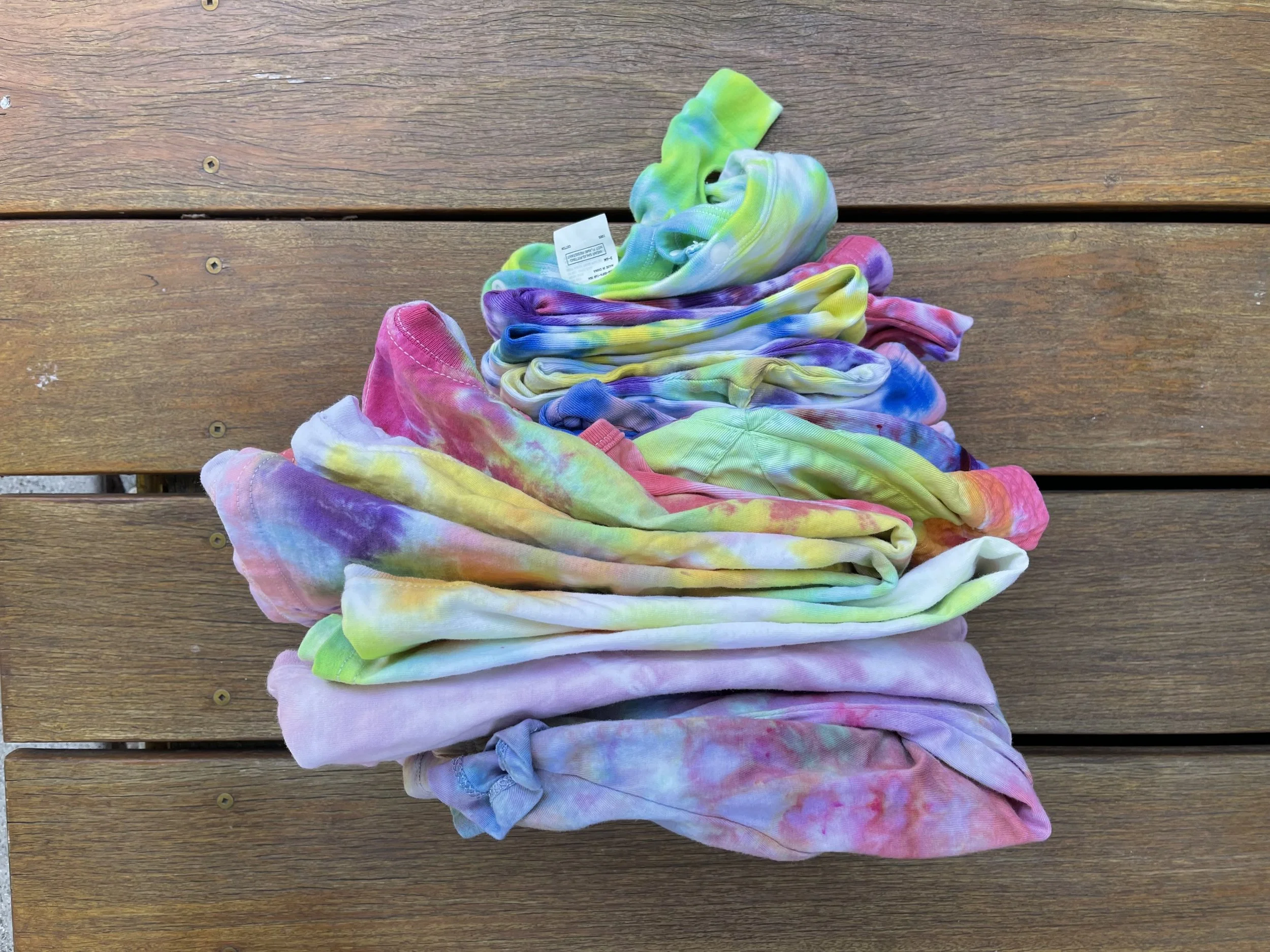 A pile of tie-dye shirts in various colors, folded and spread out on a wooden surface.