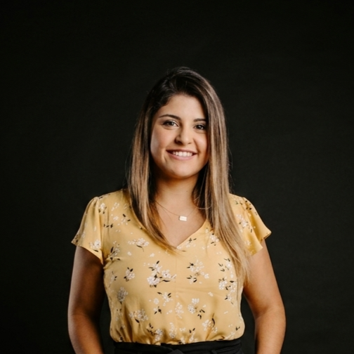 A woman wearing a yellow blazer and a white blouse standing against a black background, smiling and looking at the camera.