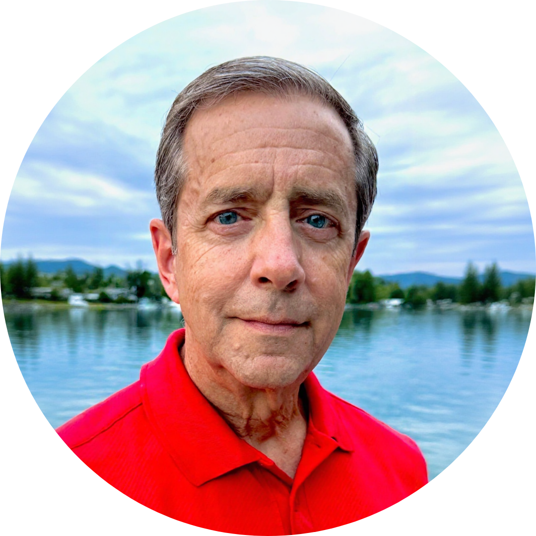 A middle-aged man with blue eyes and brown hair, wearing a red polo shirt, standing outdoors by a lake with trees and mountains in the background.