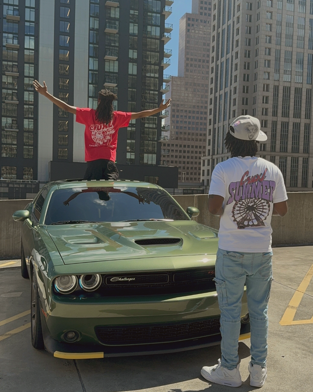 Two men in a parking garage with a green Dodge Challenger. One man is standing through the sunroof with arms outstretched, while the other man is standing behind the car wearing a white cap and a white graphic t-shirt. Tall buildings are in the backg