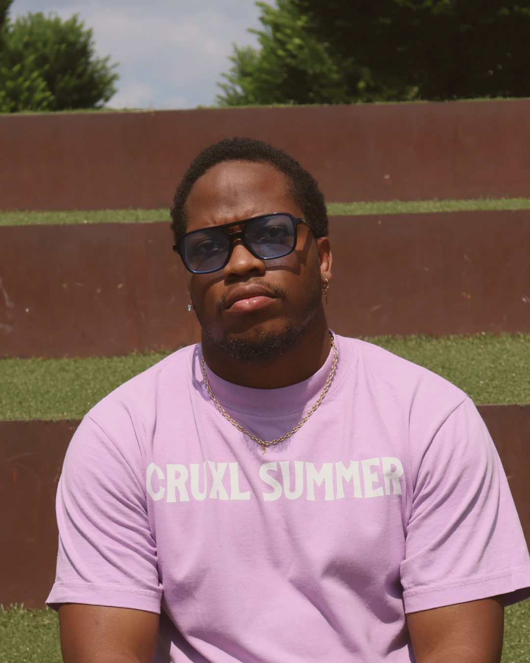 A young man with short curly hair and dark sunglasses, wearing a light pink T-shirt that says 'CRUXL SUMMER,' sitting outdoors in front of a modern wall with greenery on top, during daytime.