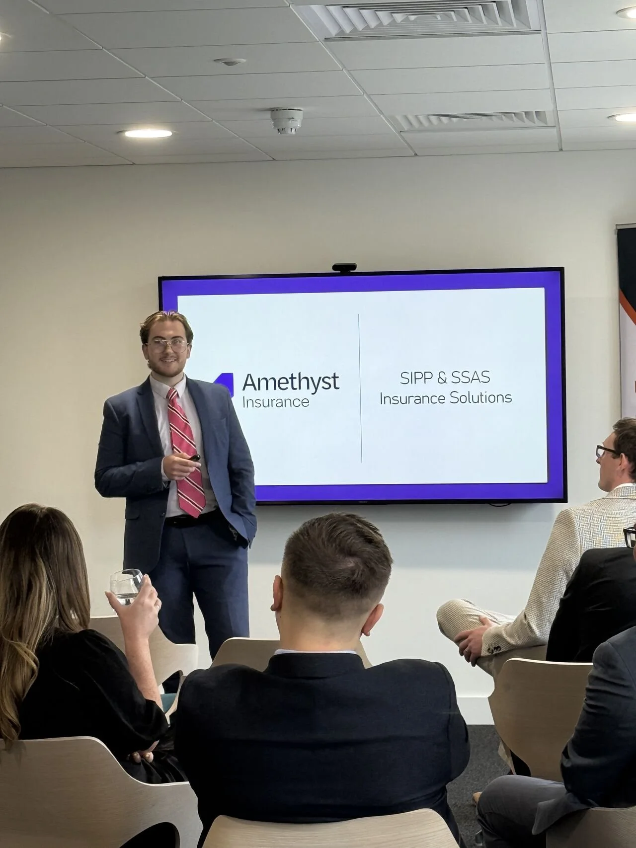 A man in a suit and pink striped tie giving a presentation in a conference room with several audience members. The slide on the screen features the company logo and the text 'Amethyst Insurance' and 'SIPP & SSAS Insurance Solutions.'