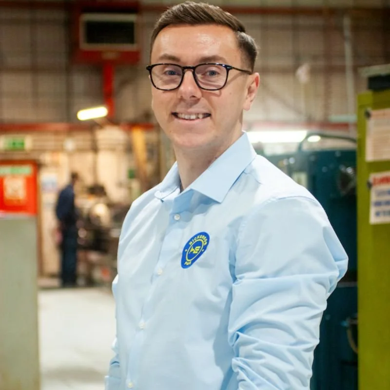 Man with glasses wearing a light blue shirt with a blue and yellow badge, standing in an industrial or warehouse setting.