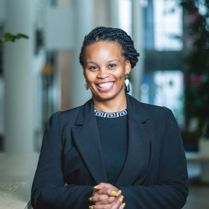 A smiling African American woman in business attire standing indoors near a window.