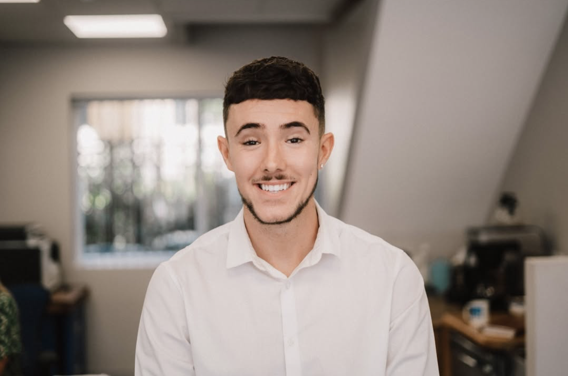 Smiling young man with short dark hair and a light beard wearing a white button-up shirt, standing in an indoor setting with a blurred office background.