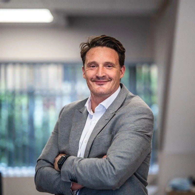 A confident man with short dark hair, wearing a light gray blazer and white shirt, standing with arms crossed in a modern office.